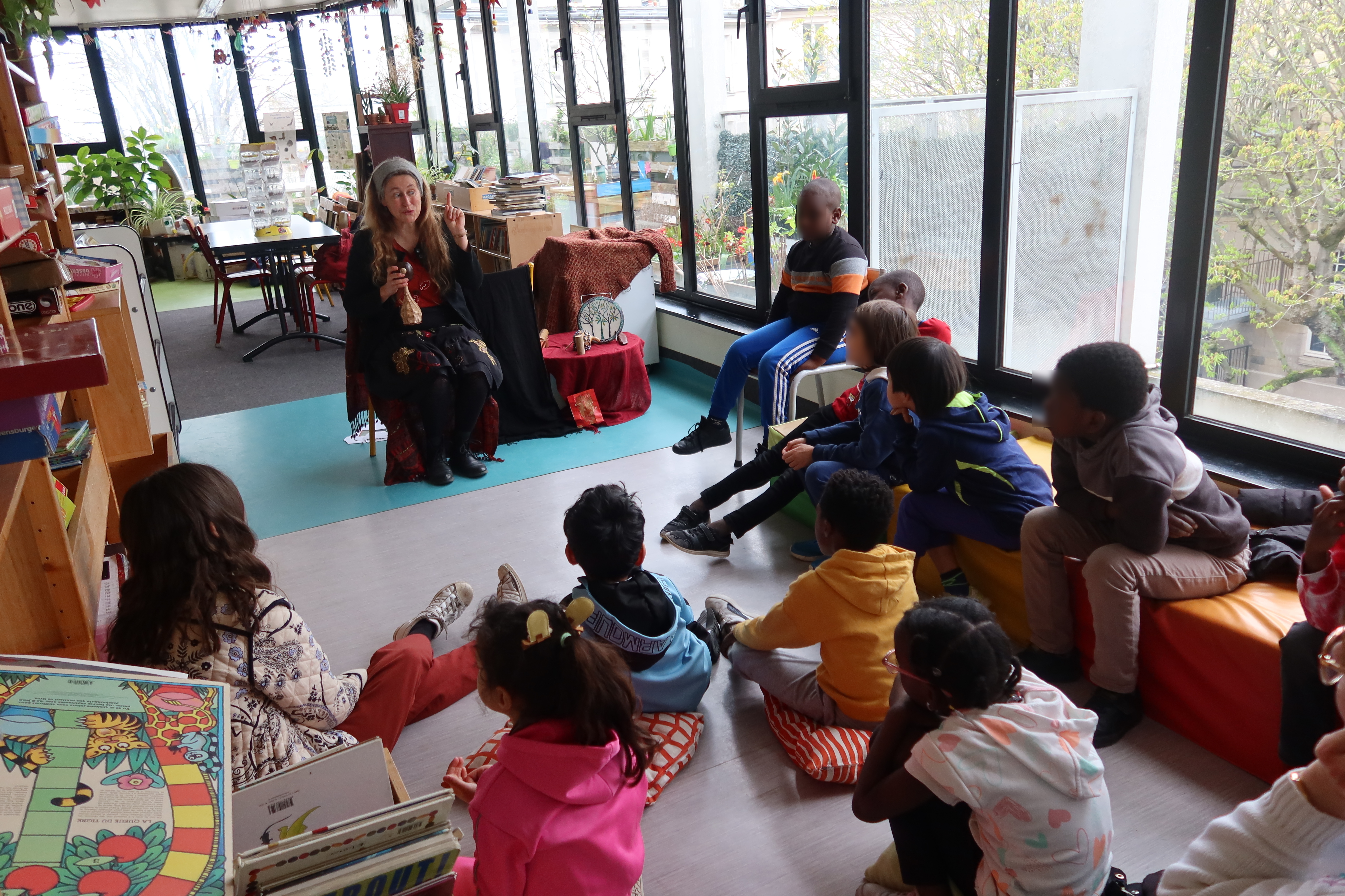 Dans la BCD de leur école, une classe est assise sur des coussins ou sur le sol. L'espace est ouvert de grandes baies vitrées. En face des élèves, assise sur une chaise, se trouve Evelyne la conteuse. A côté, d'elle, se trouve une petite table sur laquelle sont posés toutes sortes d'étranges instruments.