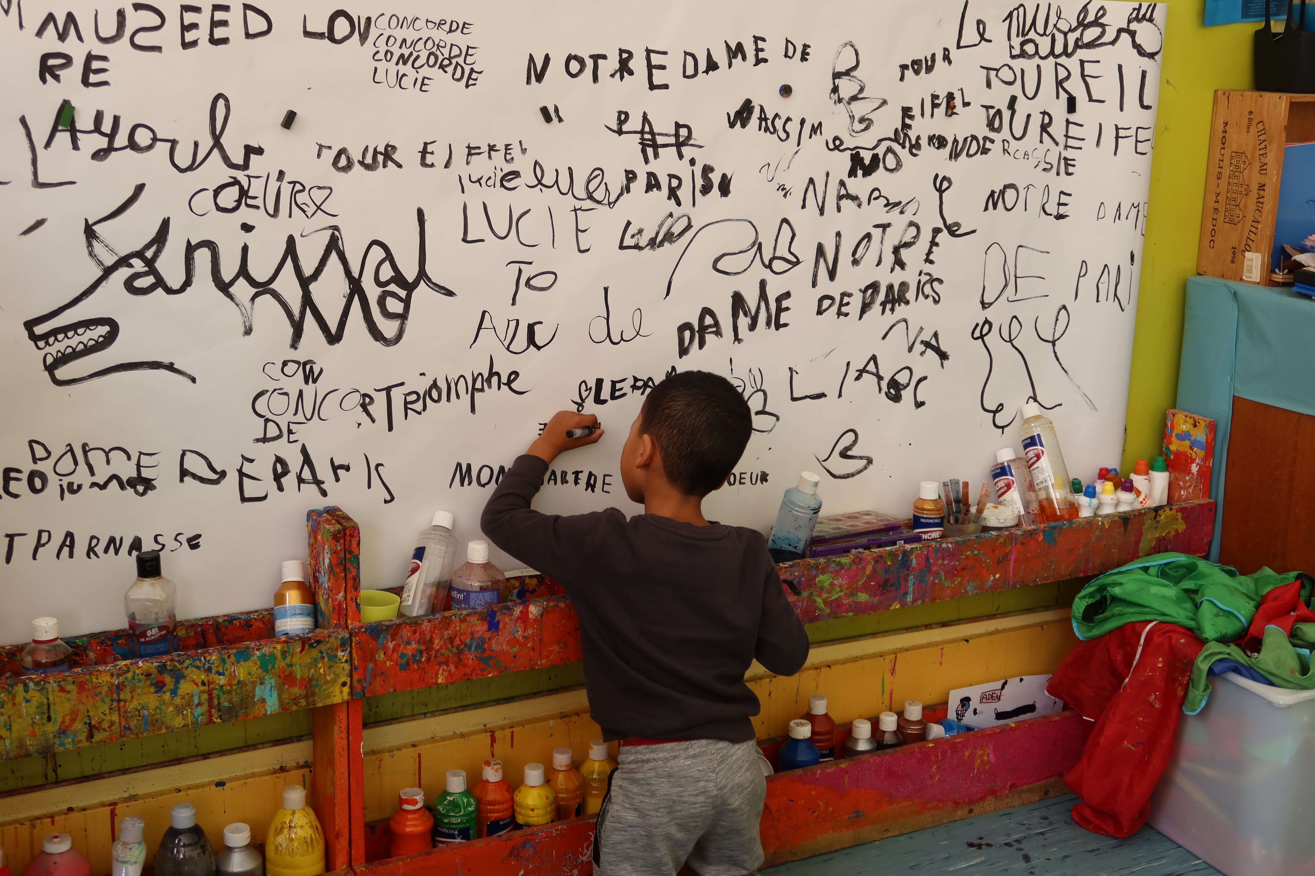 Un enfant est en train d'écrire à l'encre noir sur une grande feuilles fixée au mur qui est déjà couverte d'une multitude de mots.