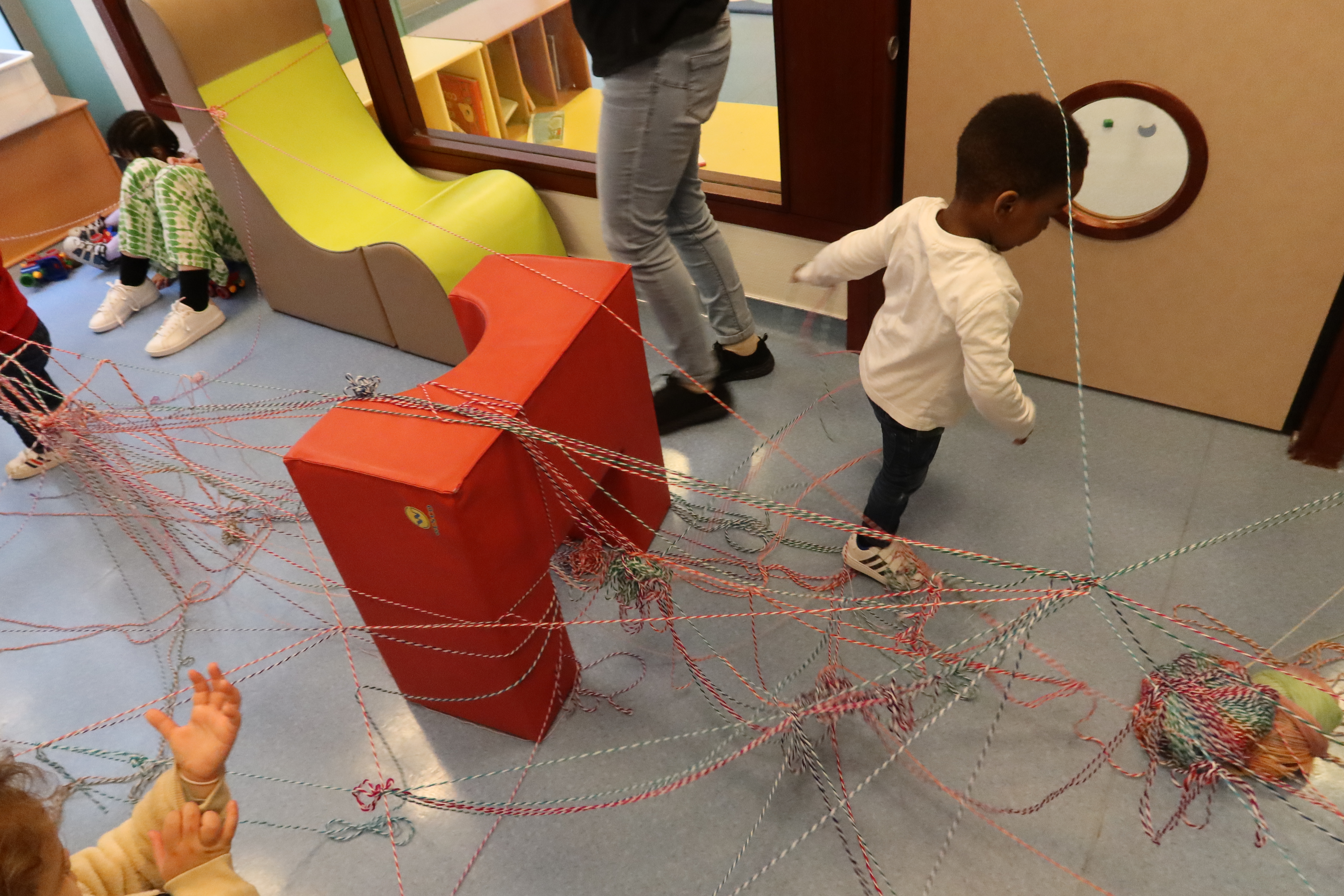 Dans une des salles de la crèche a été installée toute une toile de fils de laine et de blocs de construction en mousse pour simuler un nid. L'installation forme une sorte de parcours d’obstacle. Un petit garçon essaye de se déplacer au milieu des fils, mais il est tout emmêlé.