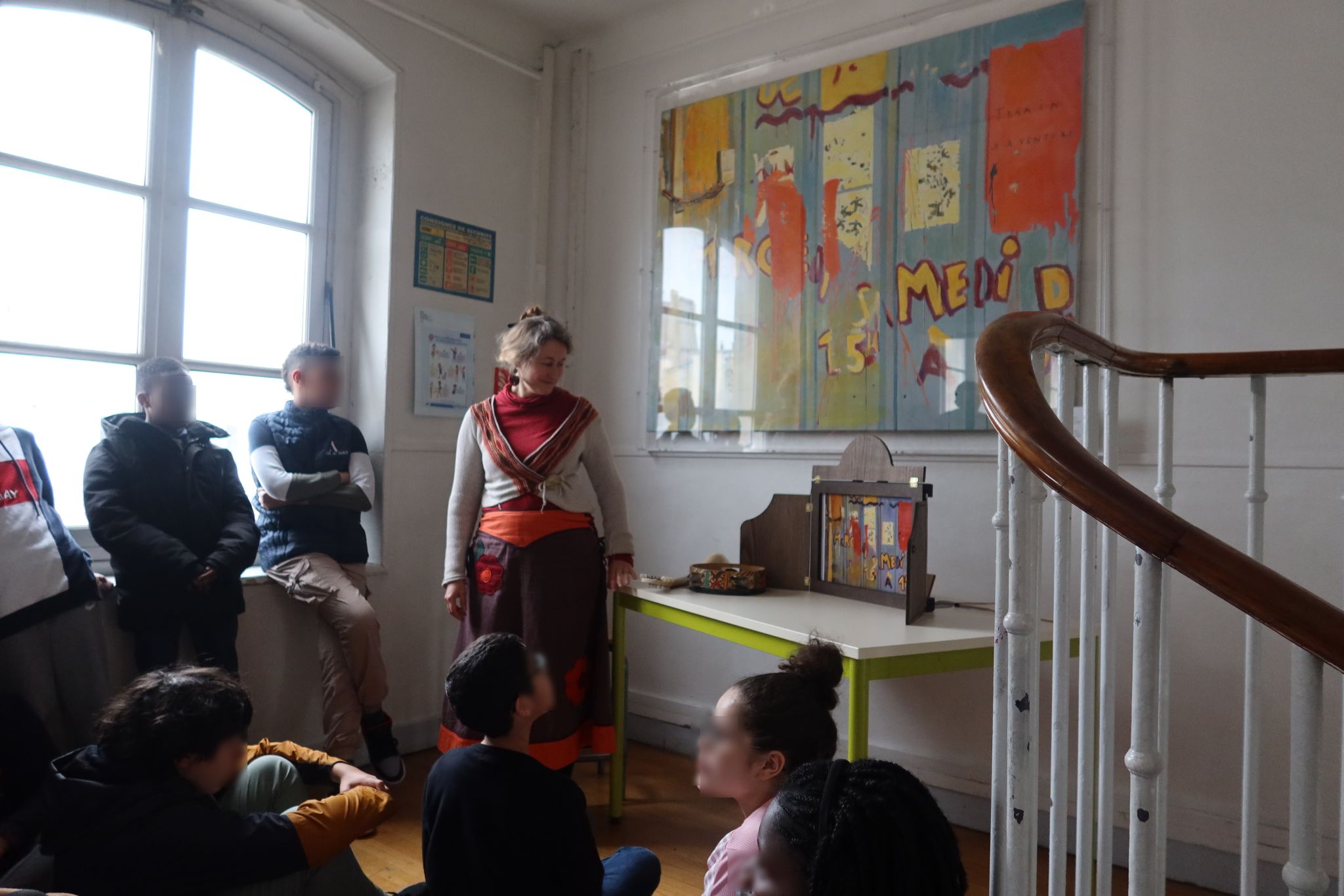 A proximité d'un grand tableau, Evelyne s'adresse aux enfants qui sont assis sur le sol du couloir ou adossés contre la fenêtre.