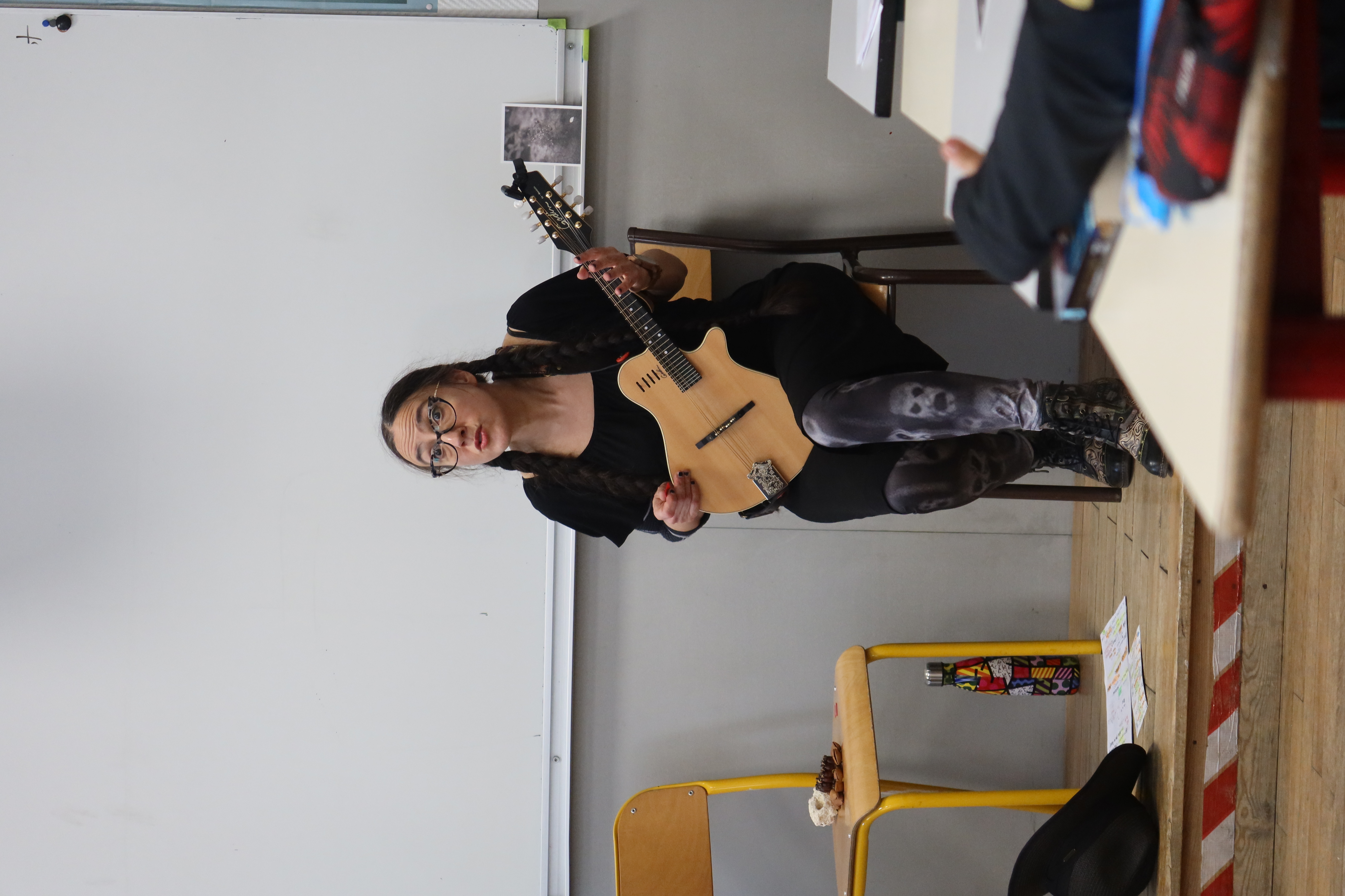 Assise sur une chaise, sur une estrade de salle de classe, Rosaline joue de sa mandoline. Les élèves l'écoute.