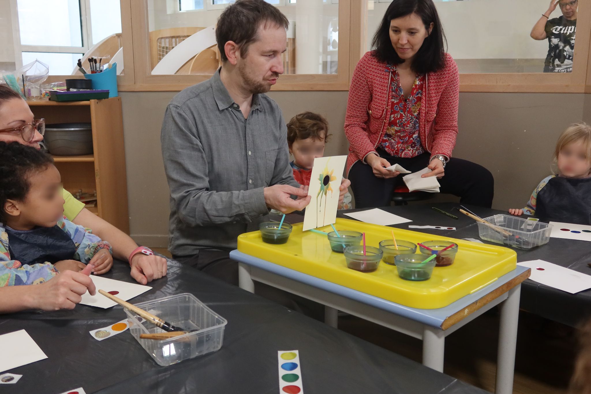 Assis autour de petites tables, les enfants, Julien Audebert, les puéricultrices et EJE ainsi que la directrice de la crèche se retrouvent pour un moment d'atelier. 
Julien Audebert vient d'expliquer en quoi l'atelier va consister et il montre maintenant la peinture qu'il a réalisé aux enfants.