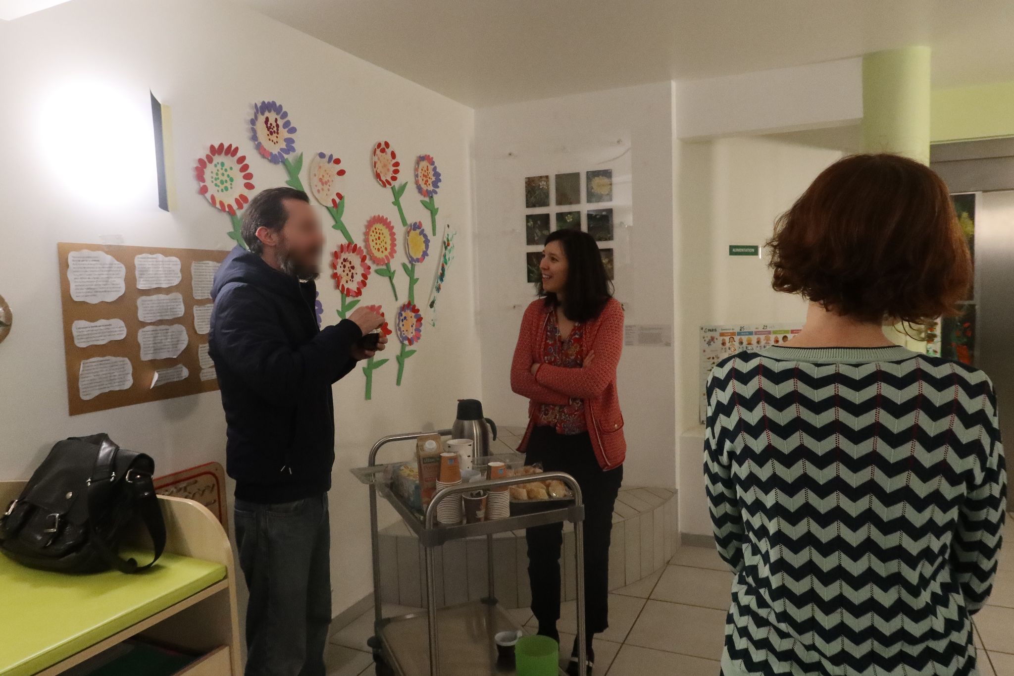 Dans le hall de la crèche, devant l'ascenseur. Les œuvres réalisées par les enfants durant les ateliers de médiation ont été exposées dans toute la pièce, sur les murs et posées sur un table. Sur un chariot ont été déposées des viennoiseries ainsi que des carafes de thé et de café. Des parents s'arrêtent boire un café pour discuter avec Julien Audebert.