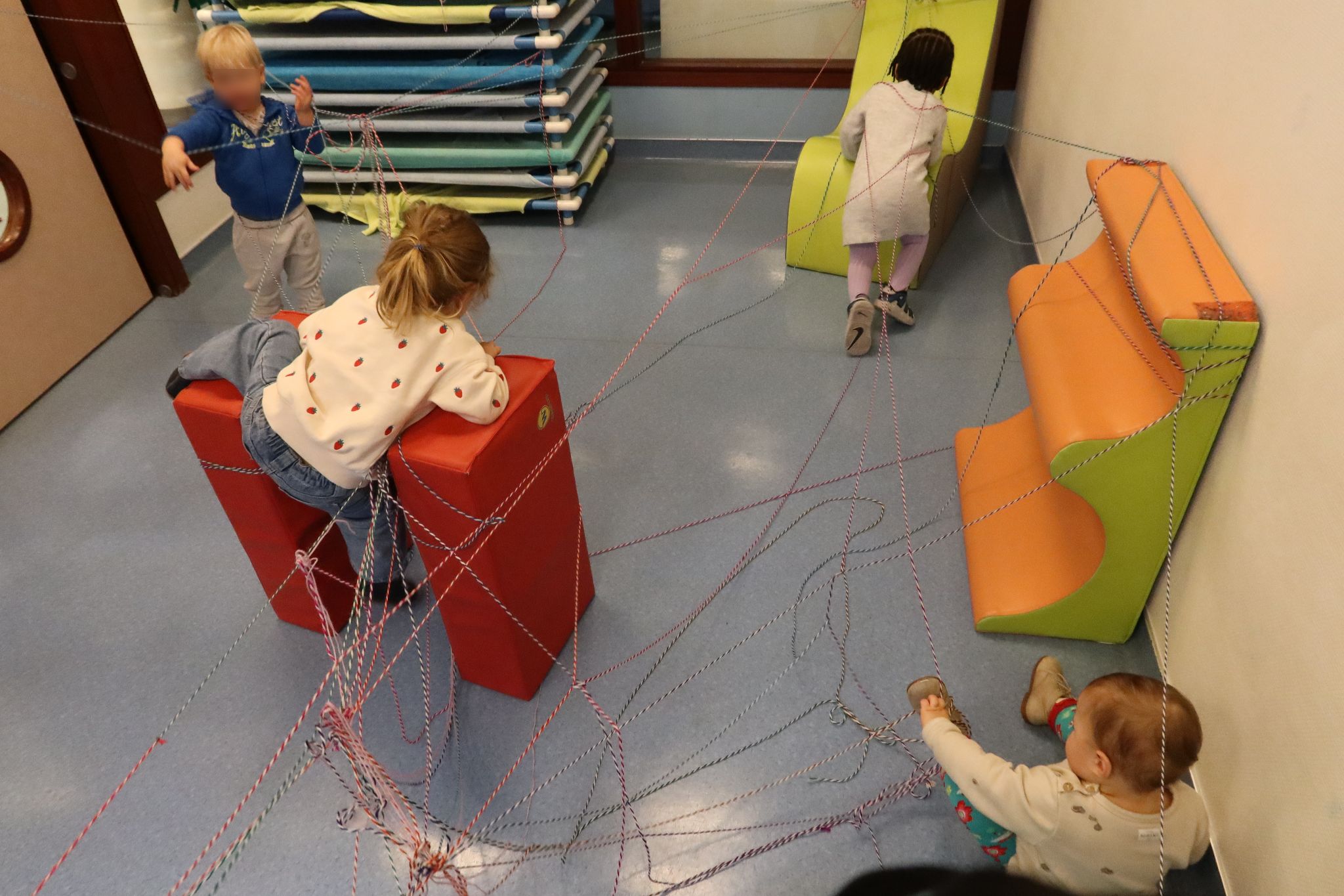 Dans une des salles de la crèche a été installée toute une toile de fils de laine et de blocs de construction en mousse pour simuler un nid. L'installation forme une sorte de parcours d’obstacle. Quatre enfants explorent le nid en se déplaçant entre les fils et en escaladant les éléments en mousse.