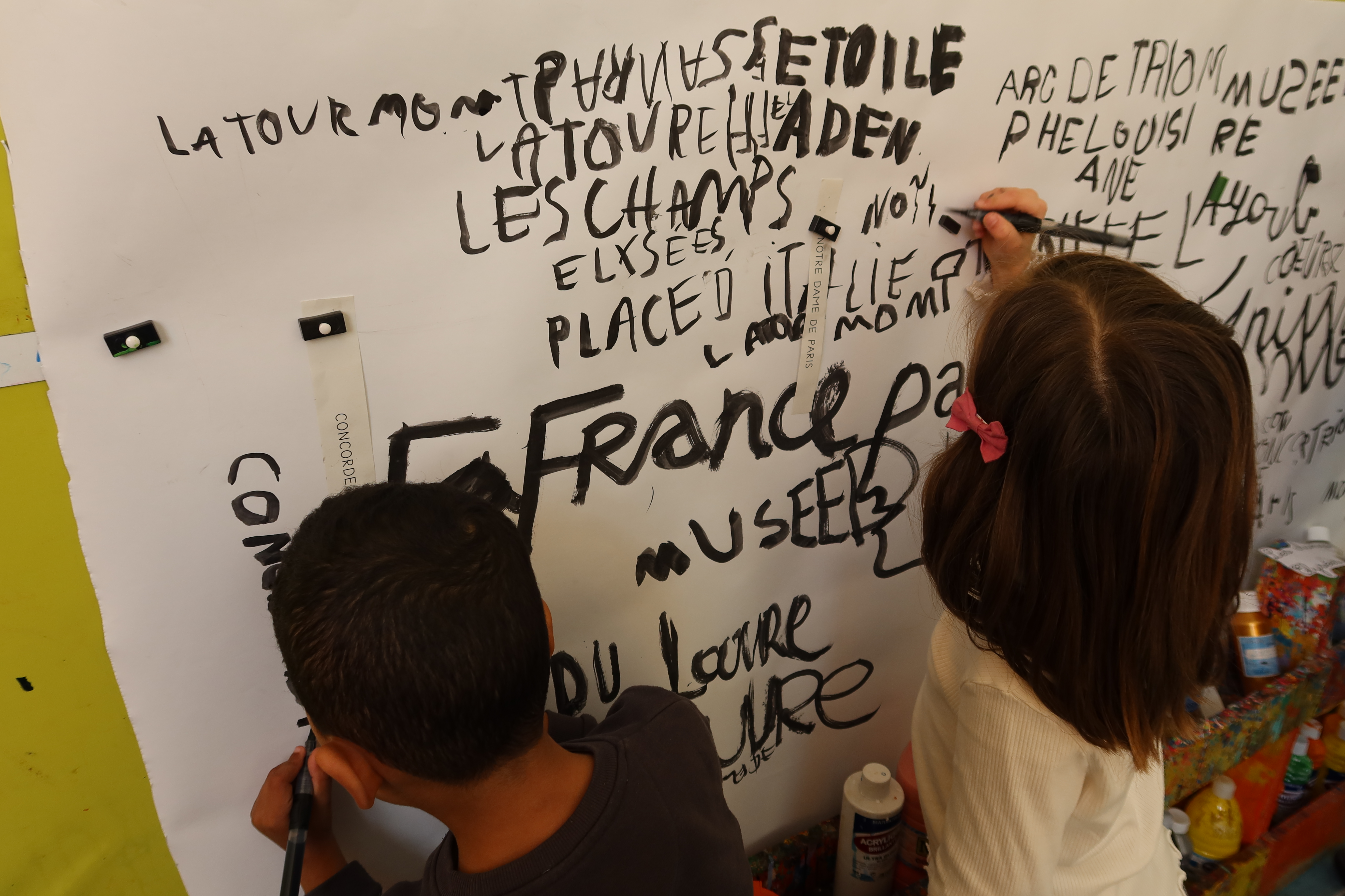 Un garçon et une fille écrive les noms des monuments français sur une feuille blanche accrochée au mur de leur salle d'art plastique
