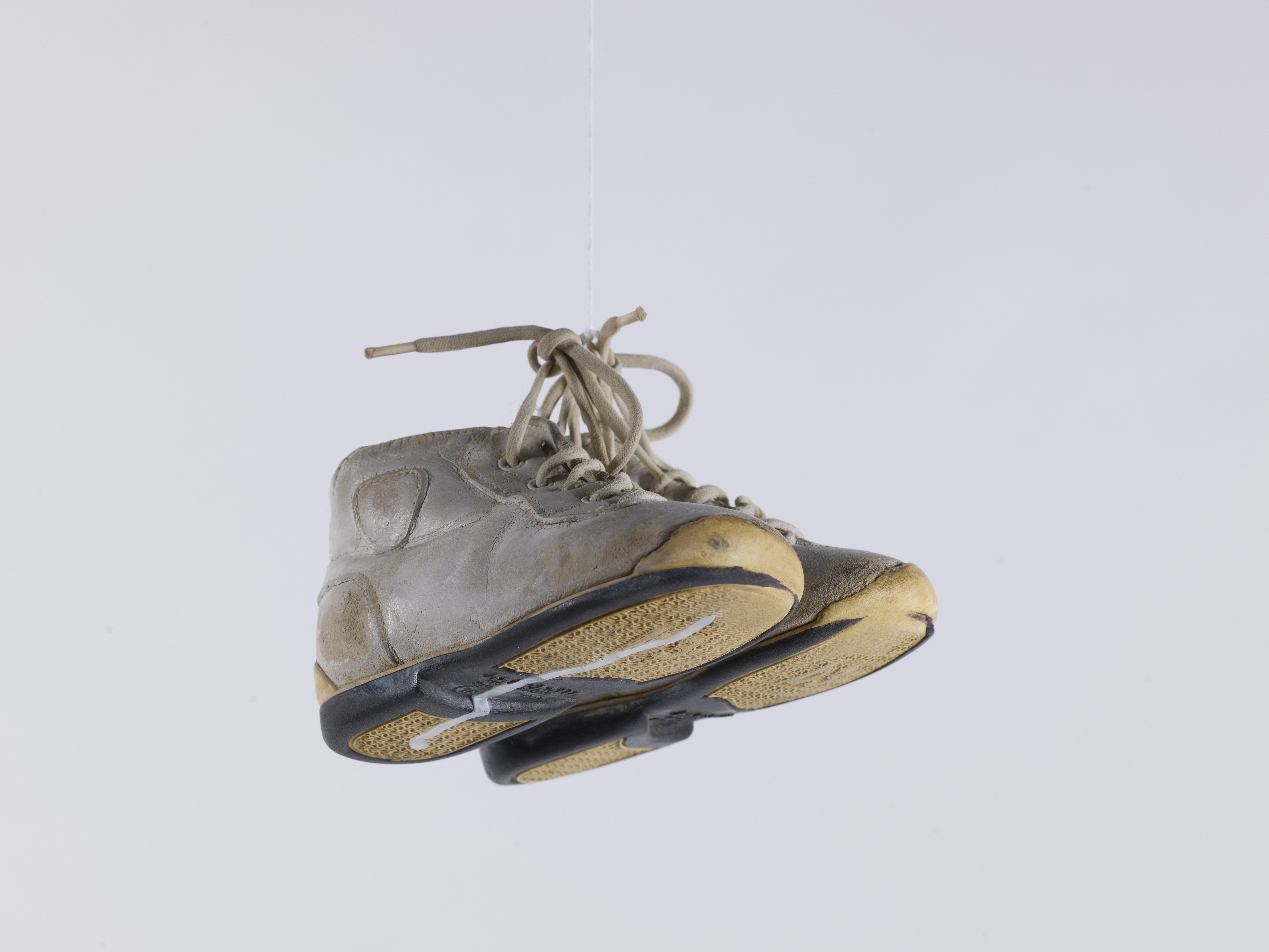 Une paire de baskets d'enfants est suspendue devant un fonds blanc par les lacets des chaussures. Sous une des semelles jaunes, on peut voir un tracé en nacre.
