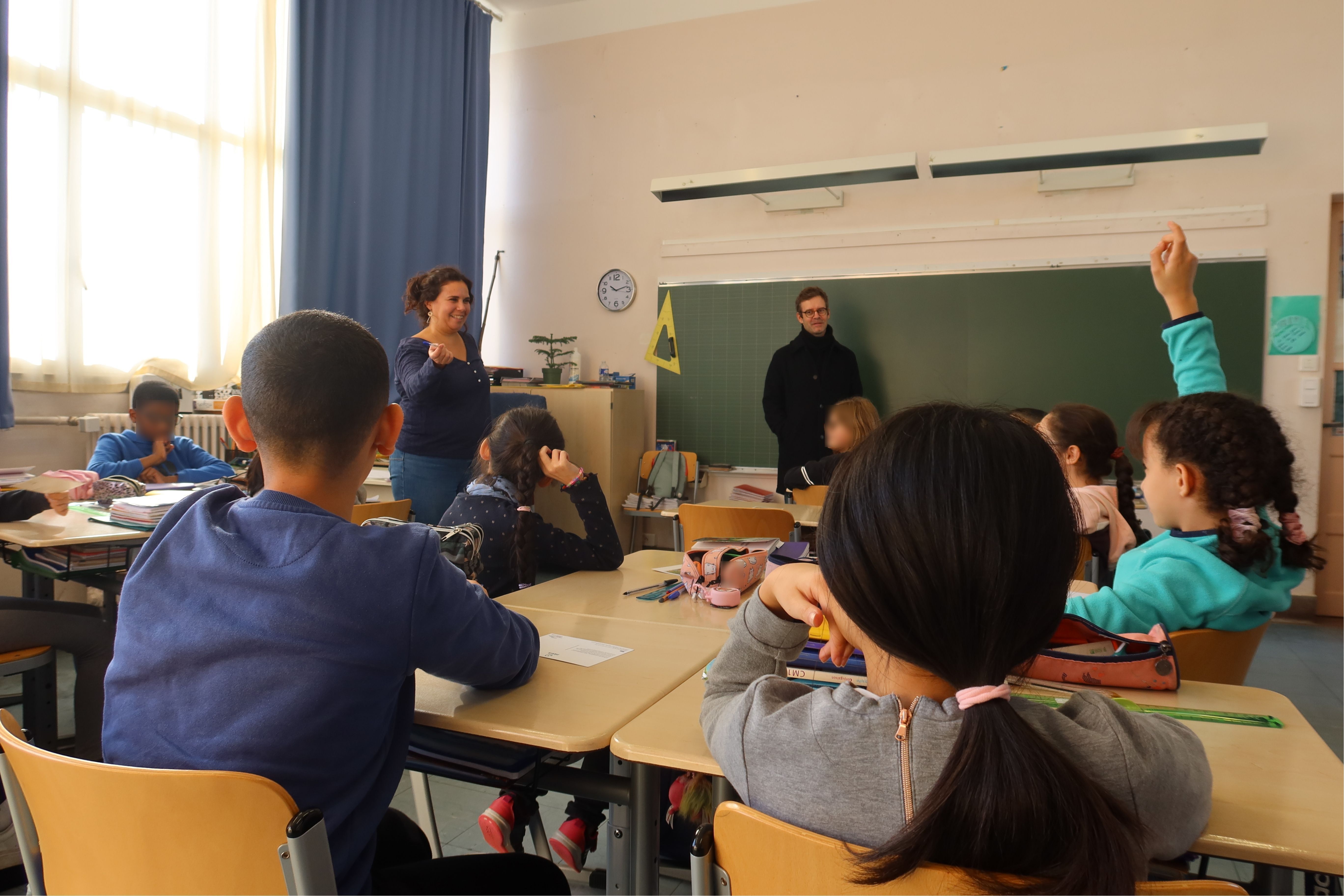 Dans une salle de classe, les élèves sont assis la main levée. Ils attendent que leur institutrice les désigne pour partager leurs impressions sur la rencontre avec Armand Jalut. L'artiste les regarde en souriant adossé au tableau noir.