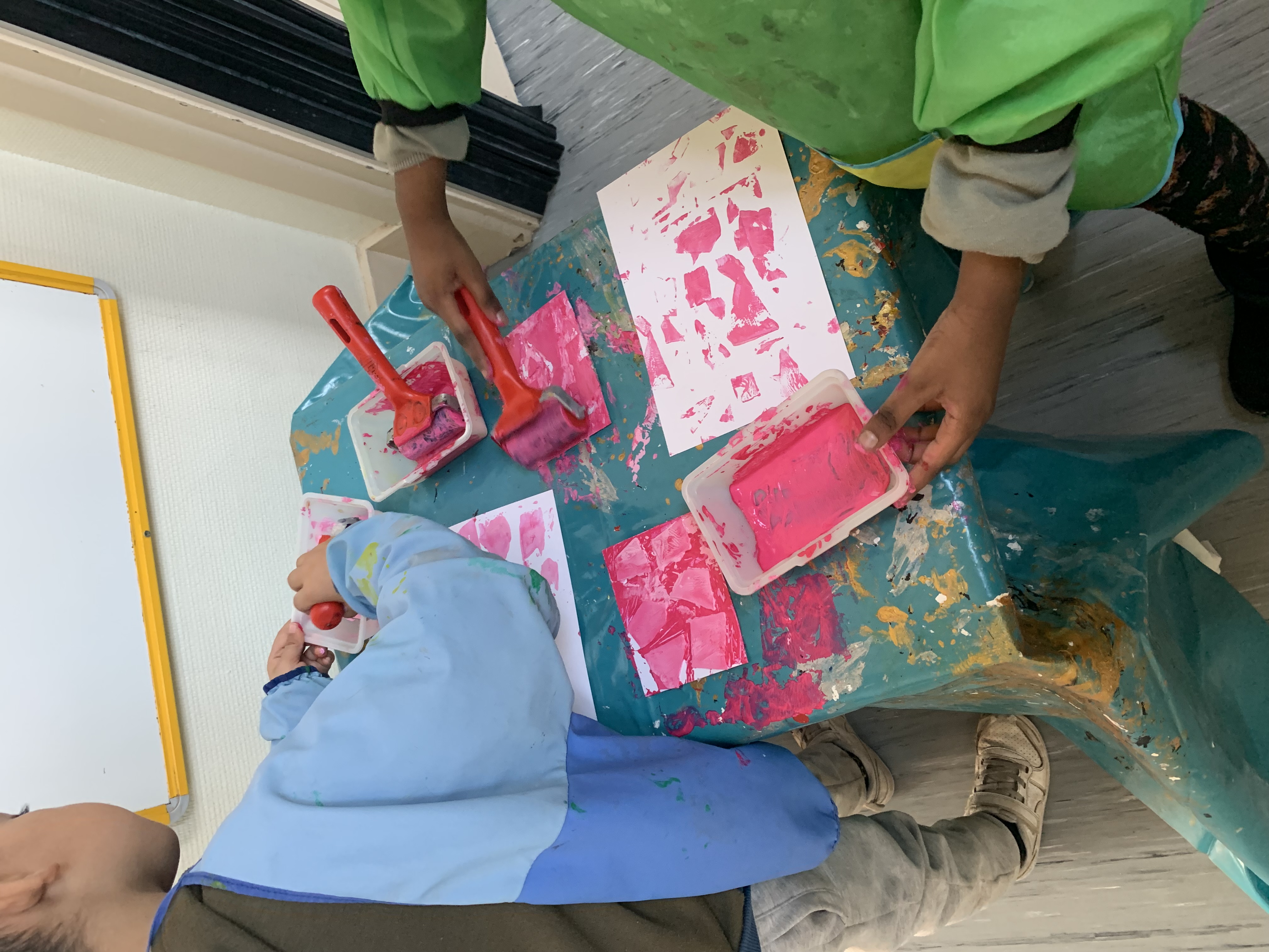 Deux enfants vêtus de tabliers de protection trempent des rouleaux en mousse dans de la peinture rose. Ils la peignent ensuite des tampons qu'ils ont fabriqué