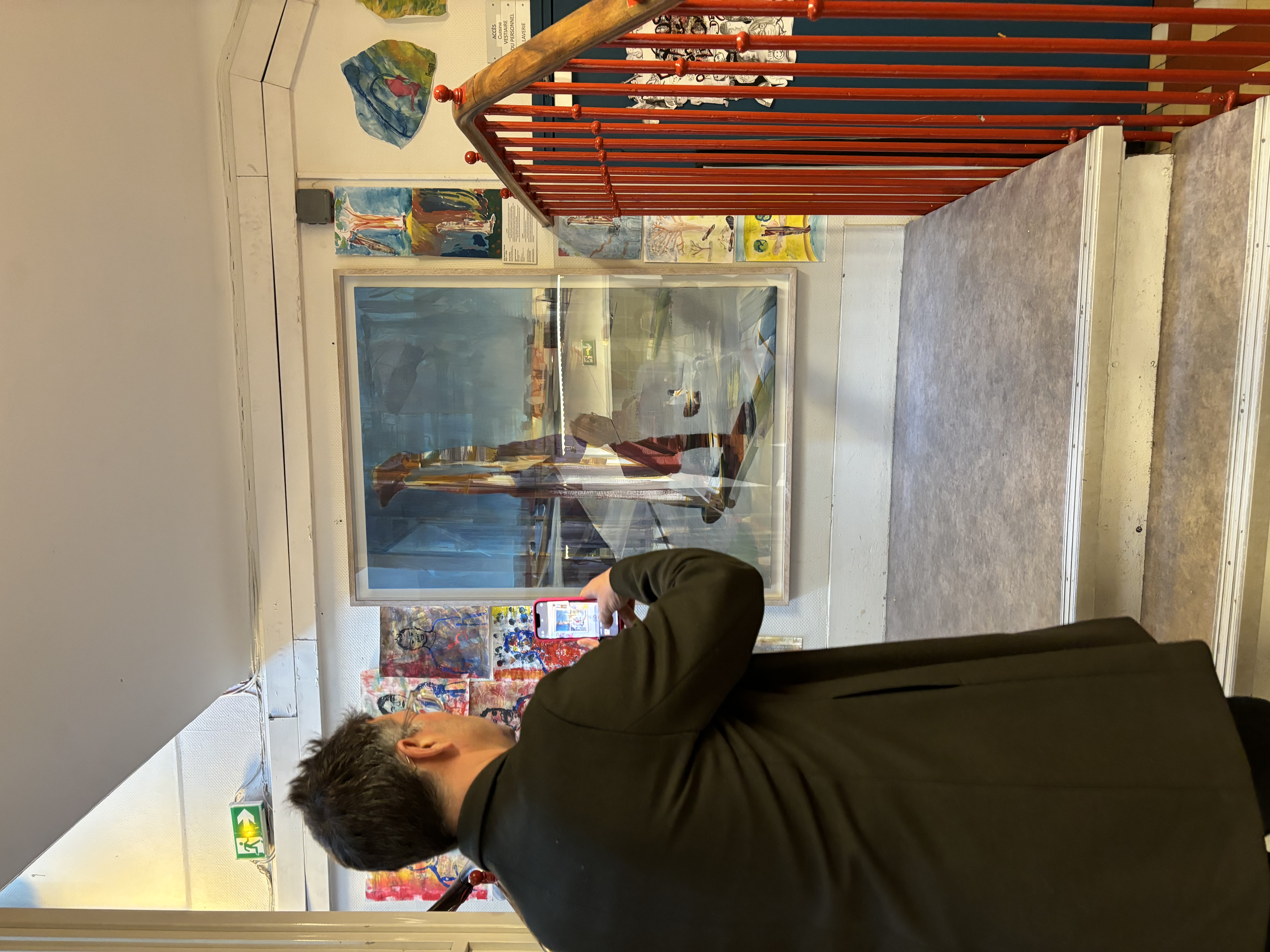 L'artiste est vêtu de noir et se tient dos à nous. Il est face à son tableau installer sur un palier d'escalier. Il est en train de prendre l’œuvre en photo avec son téléphone.