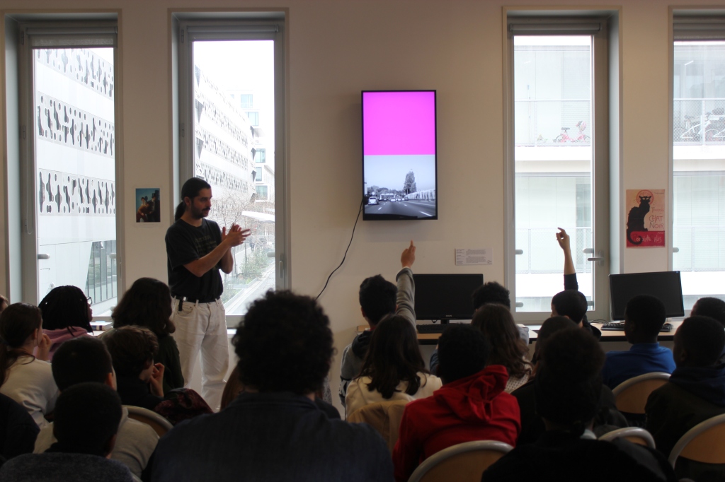 Dans une classe de collégiens, l'artiste Blaise Parmentier présente son œuvre intitulée Chromiphérie. Des enfants lèvent la main pour poser des questions.