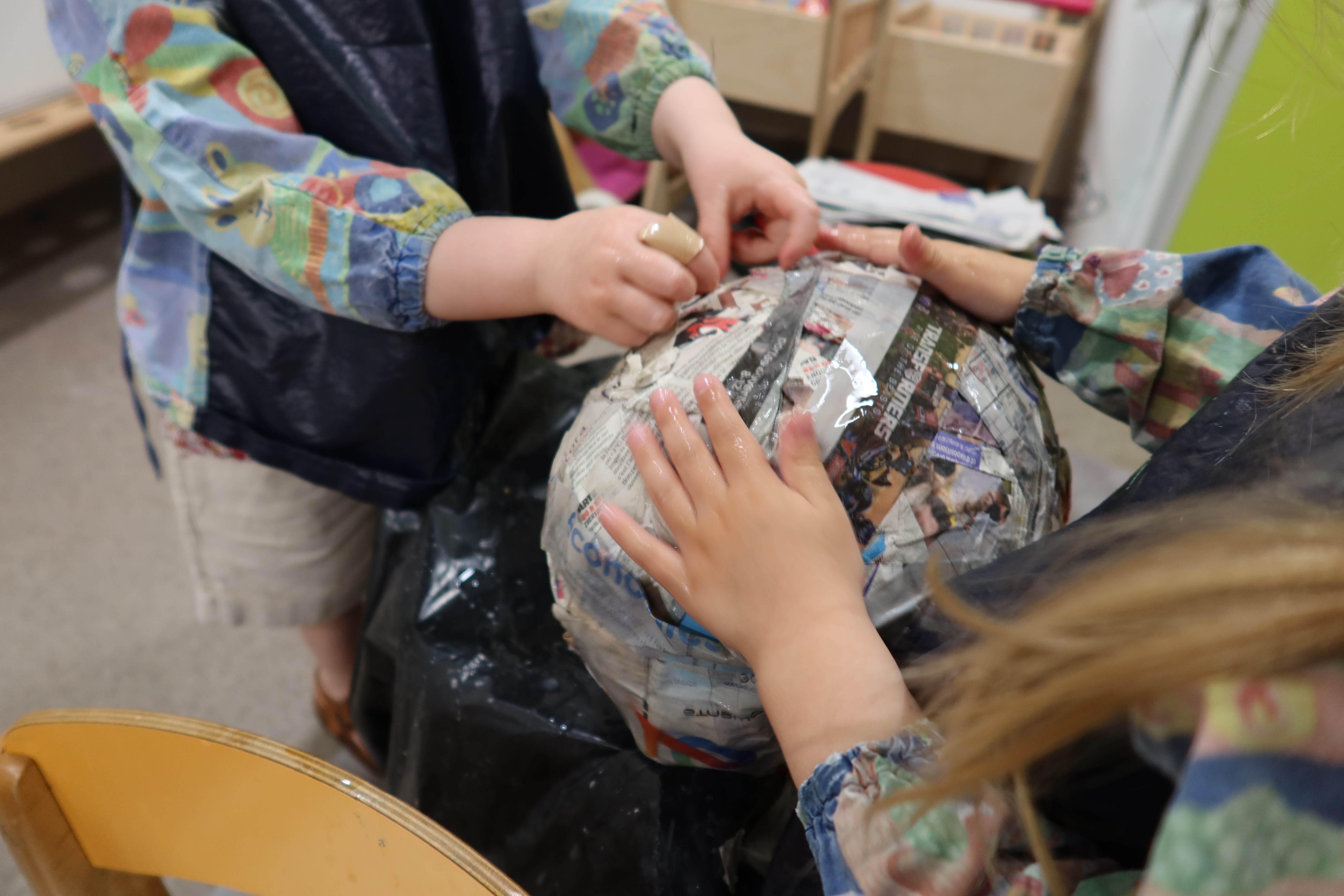 Gros plan sur les mains de deux enfants en train de coller des bandes de papier journal sur un ballon de baudruche.