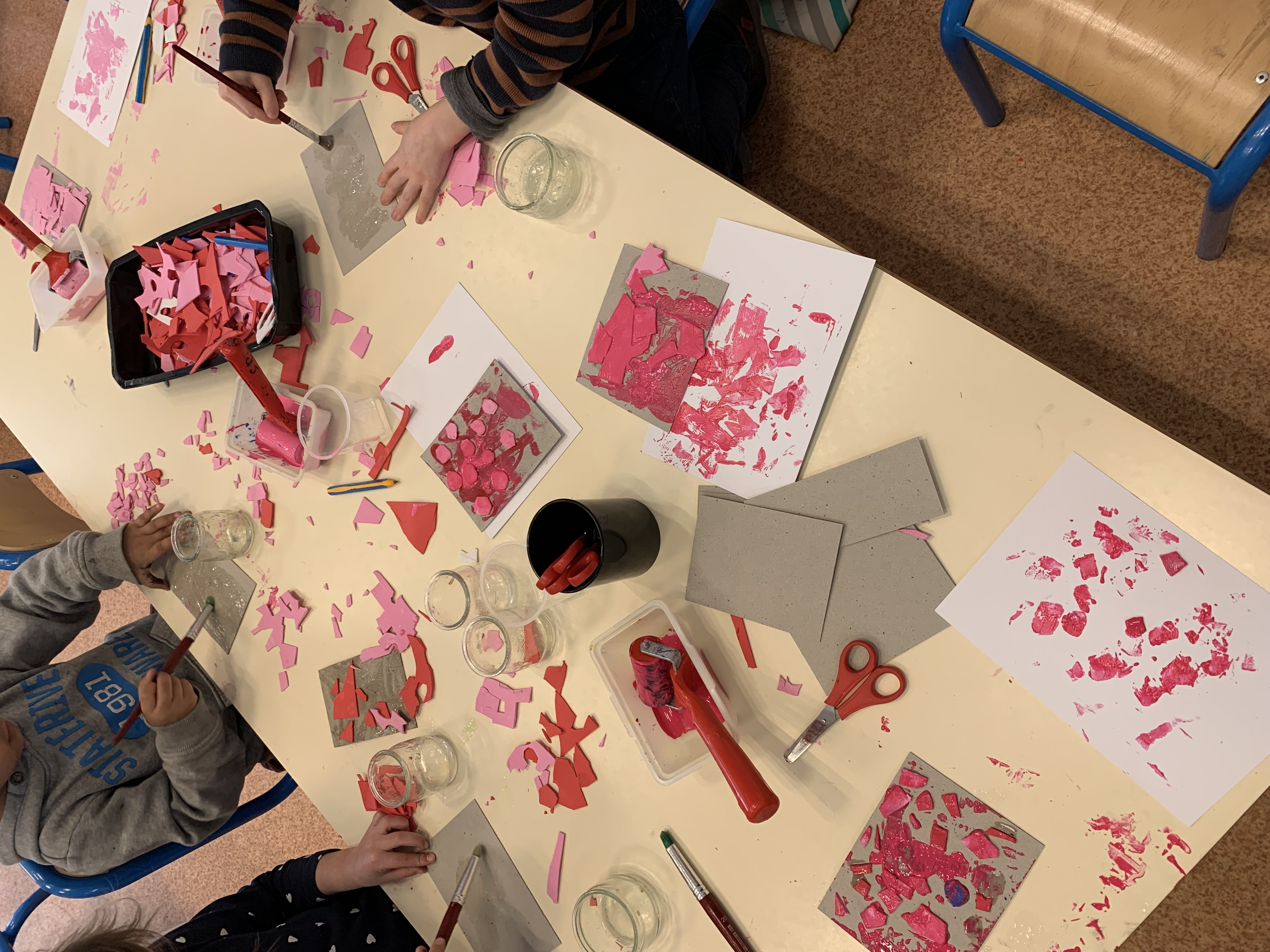 Gros plan sur une table couverte de peinture rose, de ciseaux, de feuilles de carton, de petits éléments en mousse et de pinceaux.