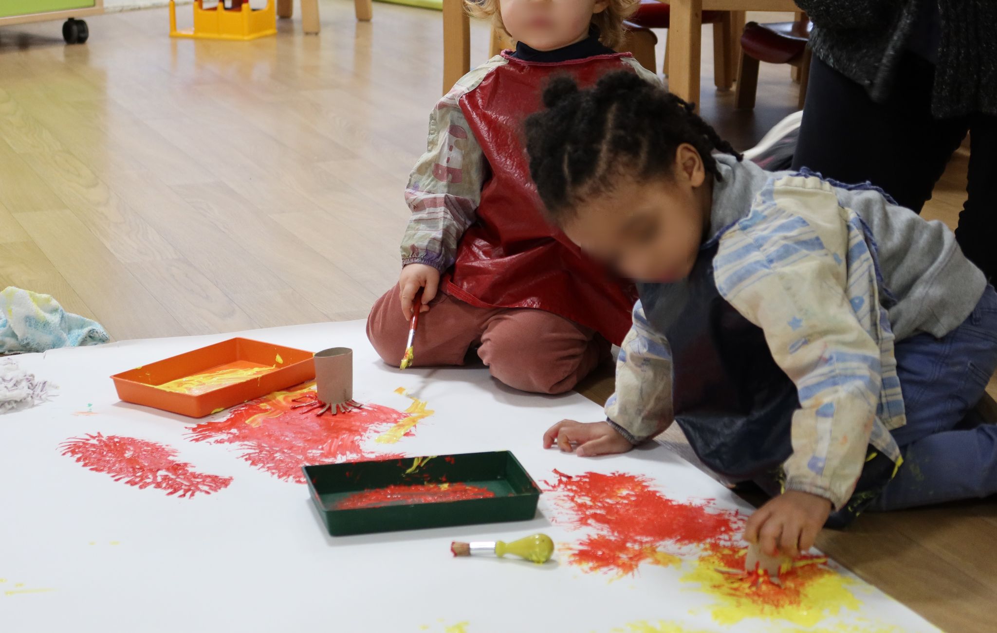 Deux enfants sont assis parterre devant une gigantesque feuille de papier. Ils sont en train de peindre à en jaune et rouge à l'aide de pinceaux et de rouleaux de PQ découpés en forme de fleurs.