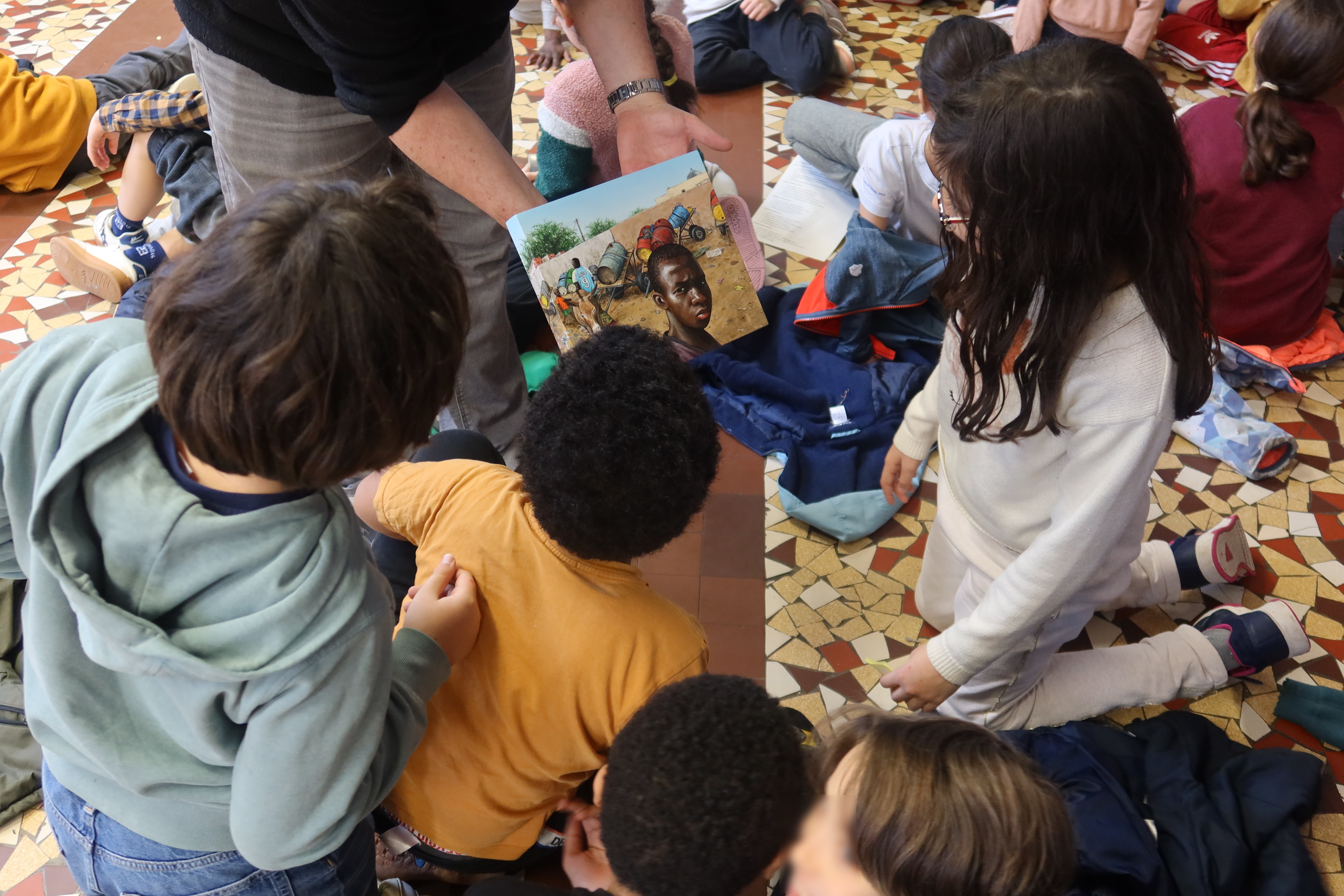 Un groupe d'enfants assis par terre se redresse pour observer de plus prêt un petit tableau qu'un adulte leur présente. Le tableau a été réalisé par Julien Beneyton lors d'un séjour au Sénégal. Il s'agit du portrait d'un garçon.