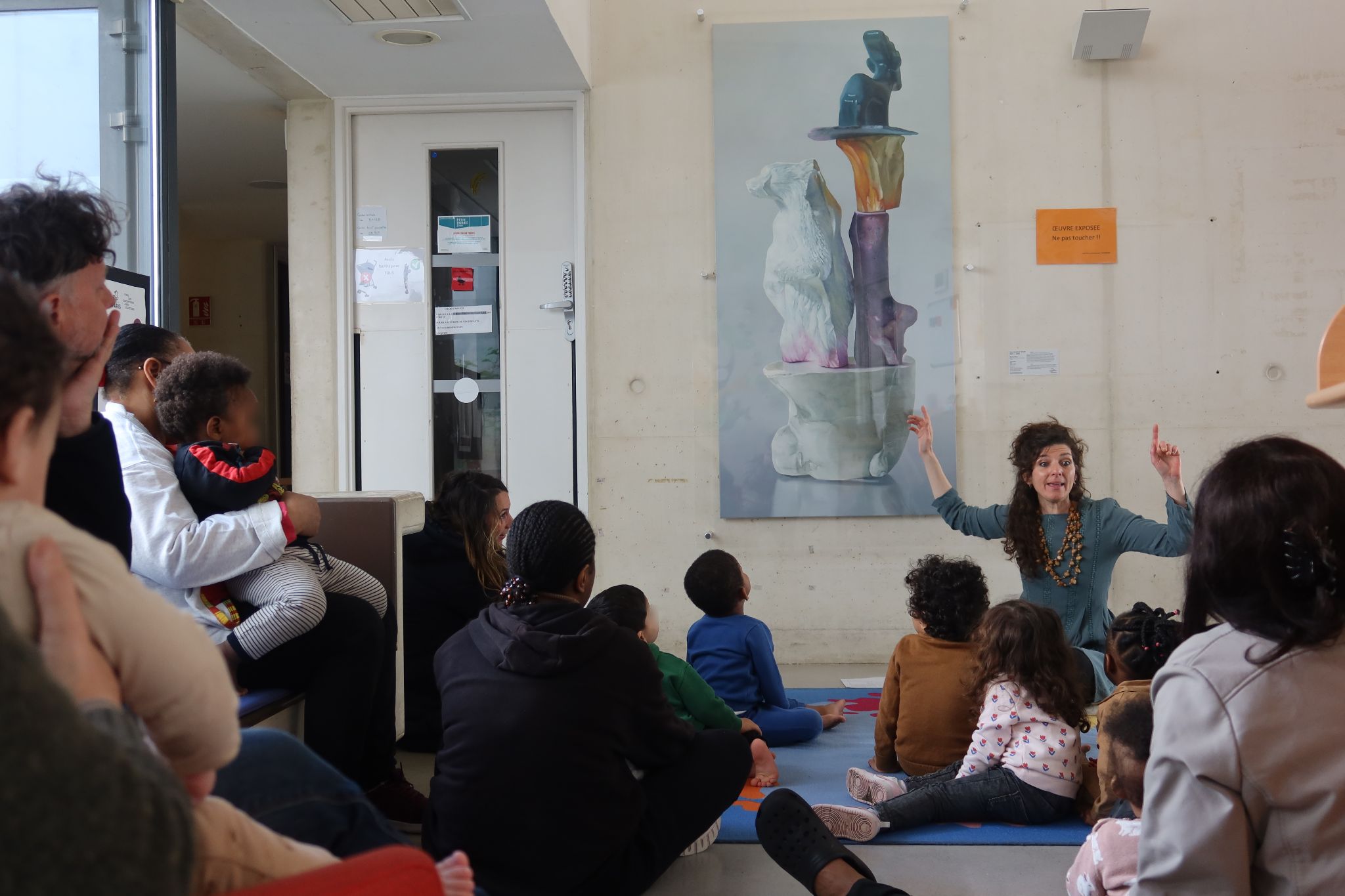 Assise devant le tableau de Maude Maris, Maïa s'adresse à un groupe d'enfants et d'adultes. Elle lève les bras pour animer l'histoire qu'elle raconte. Tout le monde la regarde attentivement.