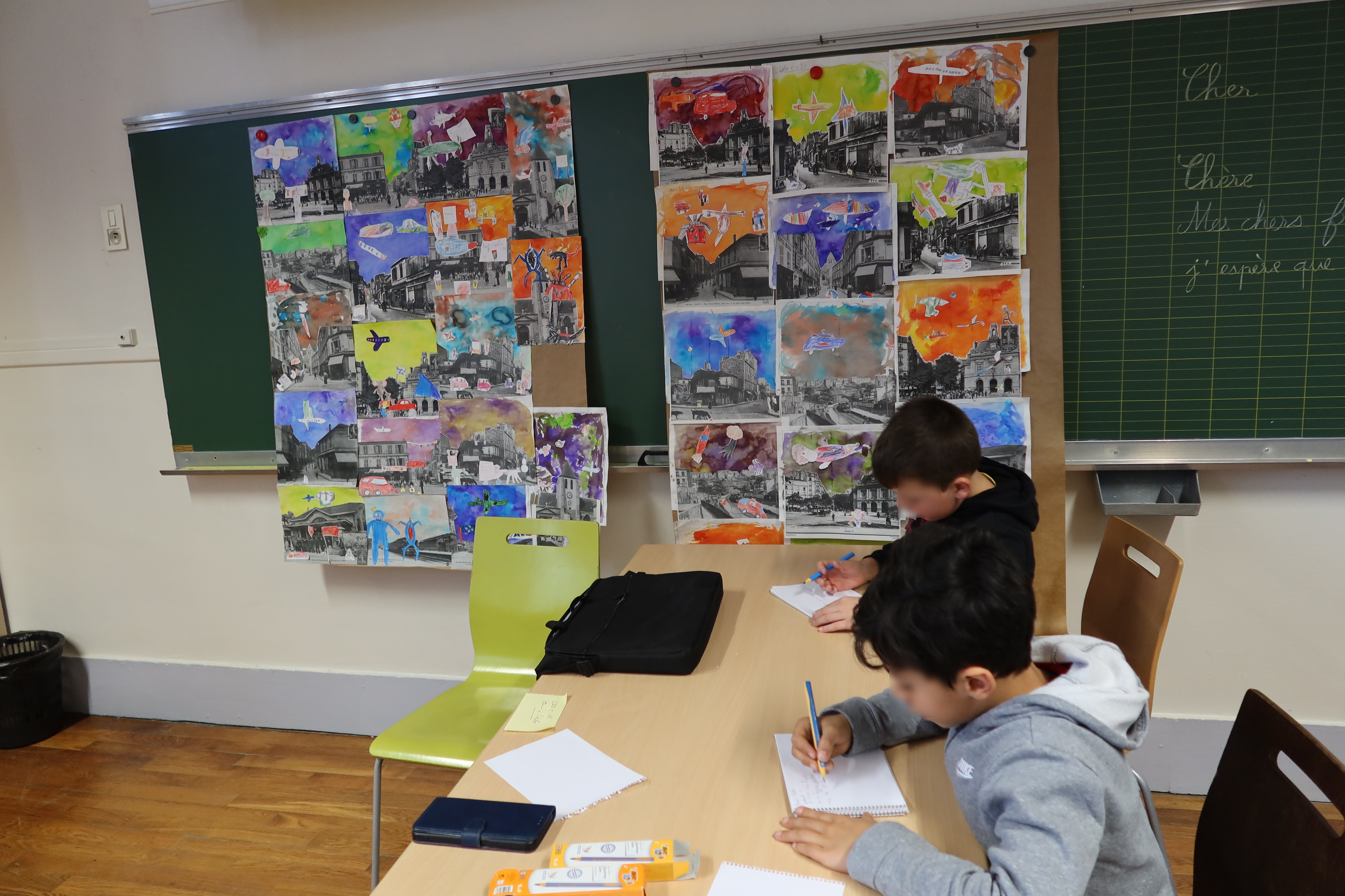 Deux élèves sont en train d'écrire leurs cartes postales. Ils sont installés à la perpendiculaire du tableau noir. Sur ce dernier se trouve affichées les aquarelles réalisées par les CE2.