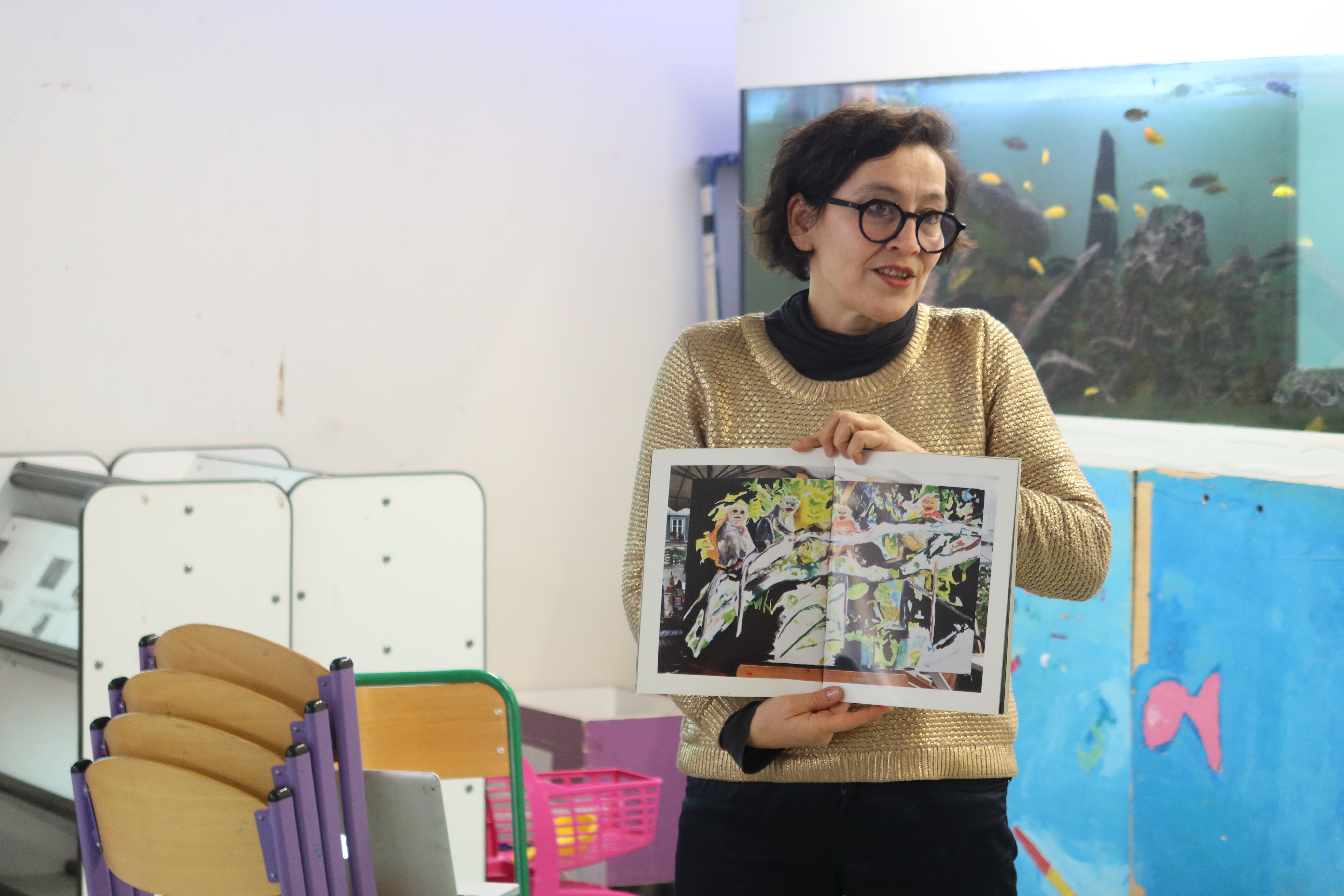 L'artiste, une femme brune avec des lunettes, présente un livre aux élèves. Le livre en grand ouvert, image vers ses interlocuteurs.