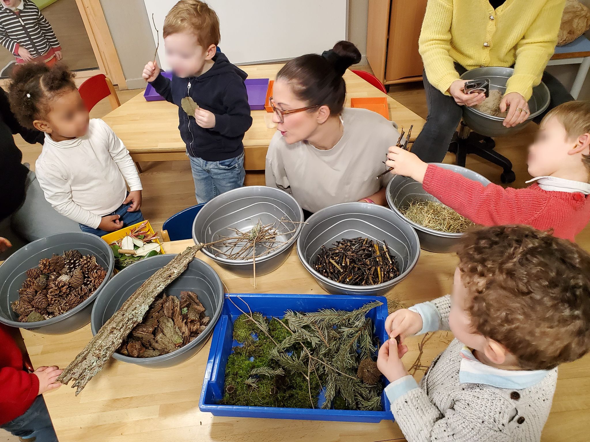 Une femme et un groupe d'enfants en bas âges sont installés autour d'une table. Sur celle-ci sont posées plusieurs bassines en plastiques remplies d'éléments naturels (écorce, pommes de pin, paille, branches de sapins, brindilles, etc.)