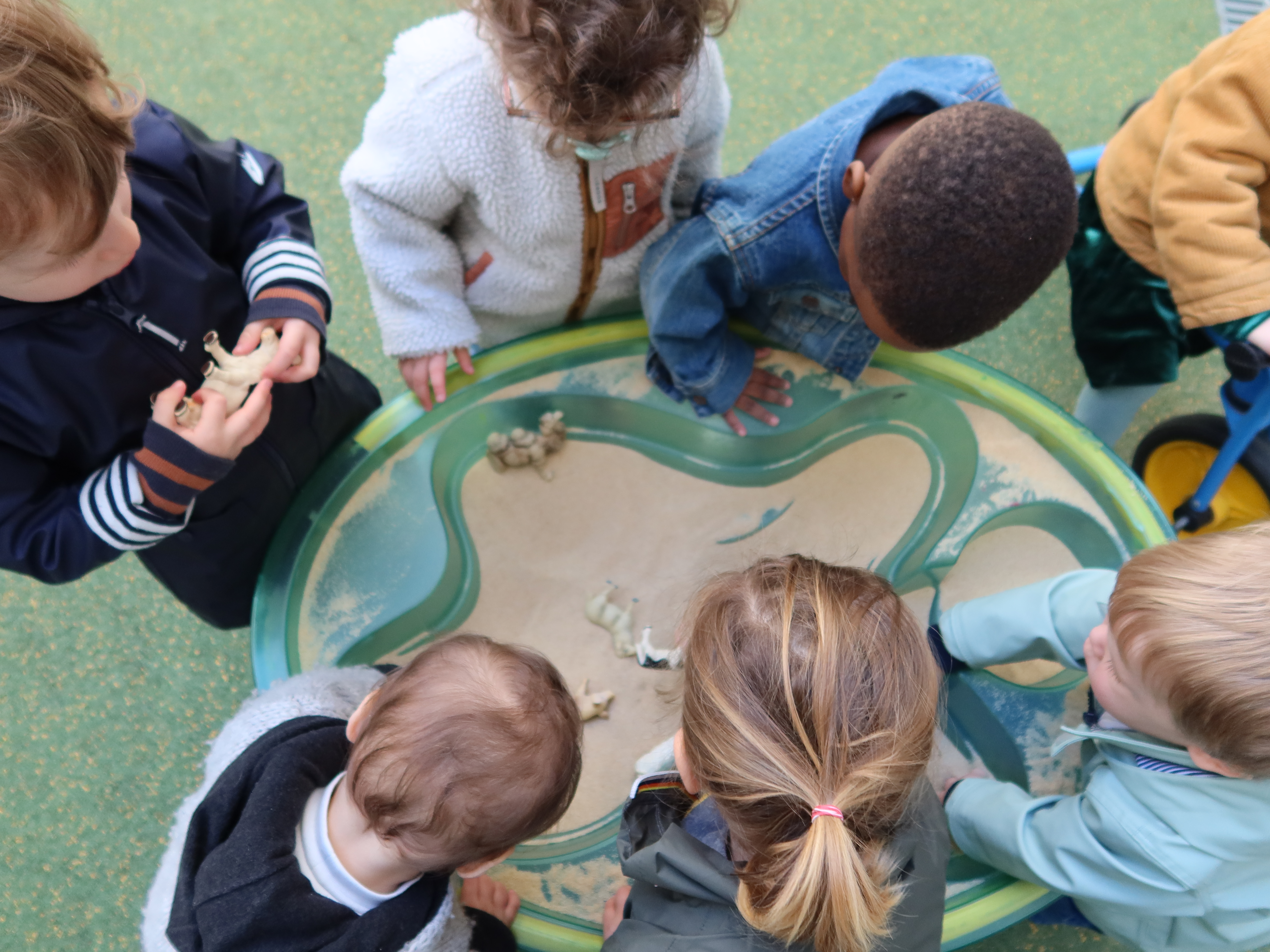 Sept enfants sont regroupés autour d'un petit bac à sable sur pied. Dans le sable sont cachés plusieurs figurines d'animaux. Le top départ a été donné, ils doivent retrouver l'un des animaux.