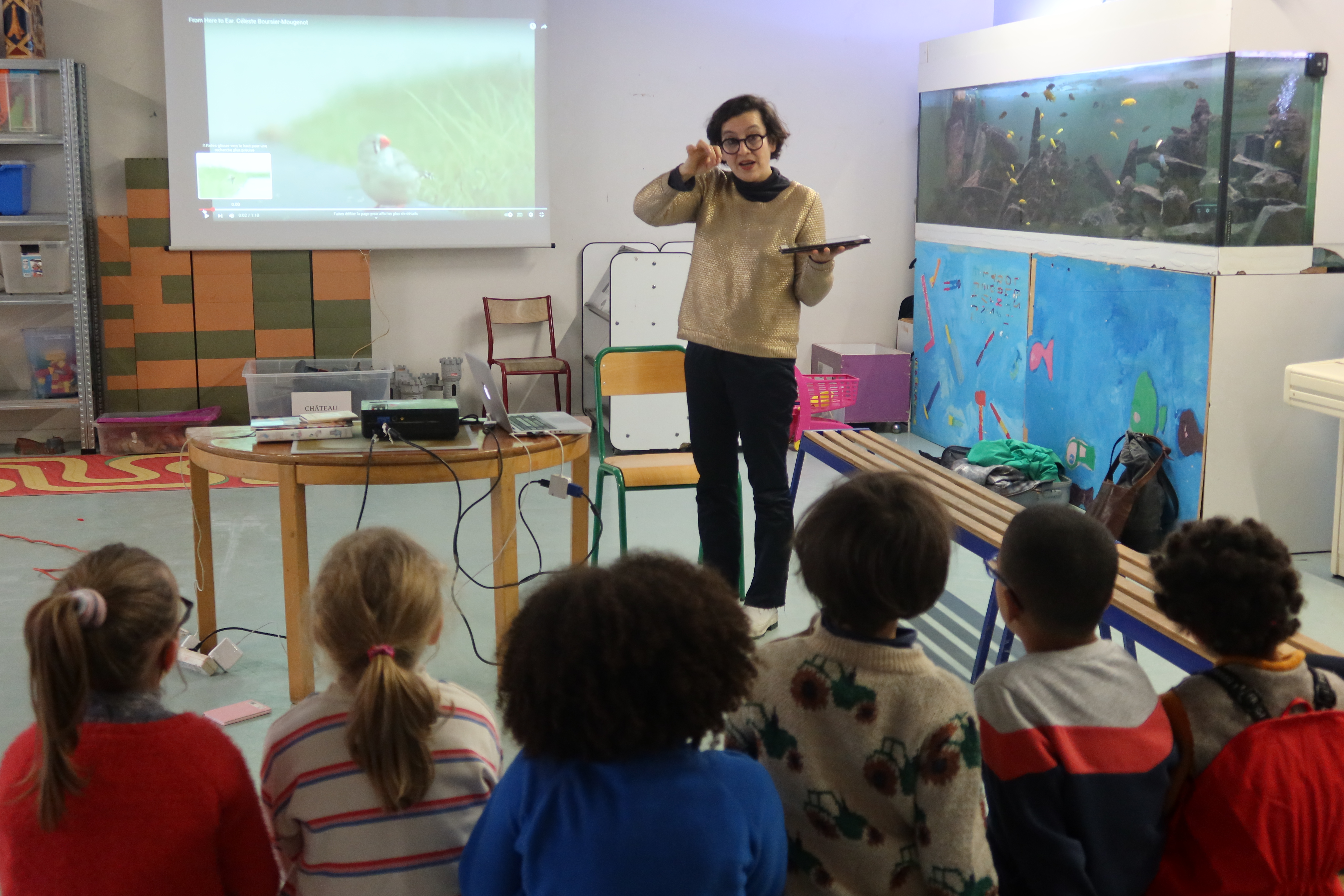 Fabienne Audéoud se tient face à un groupe d'enfants assis sur un banc. Elle tient une tablette à la main et est en train de remonter ses lunettes. Derrière elle est projetée sur un écran la vidéo d'une œuvre.