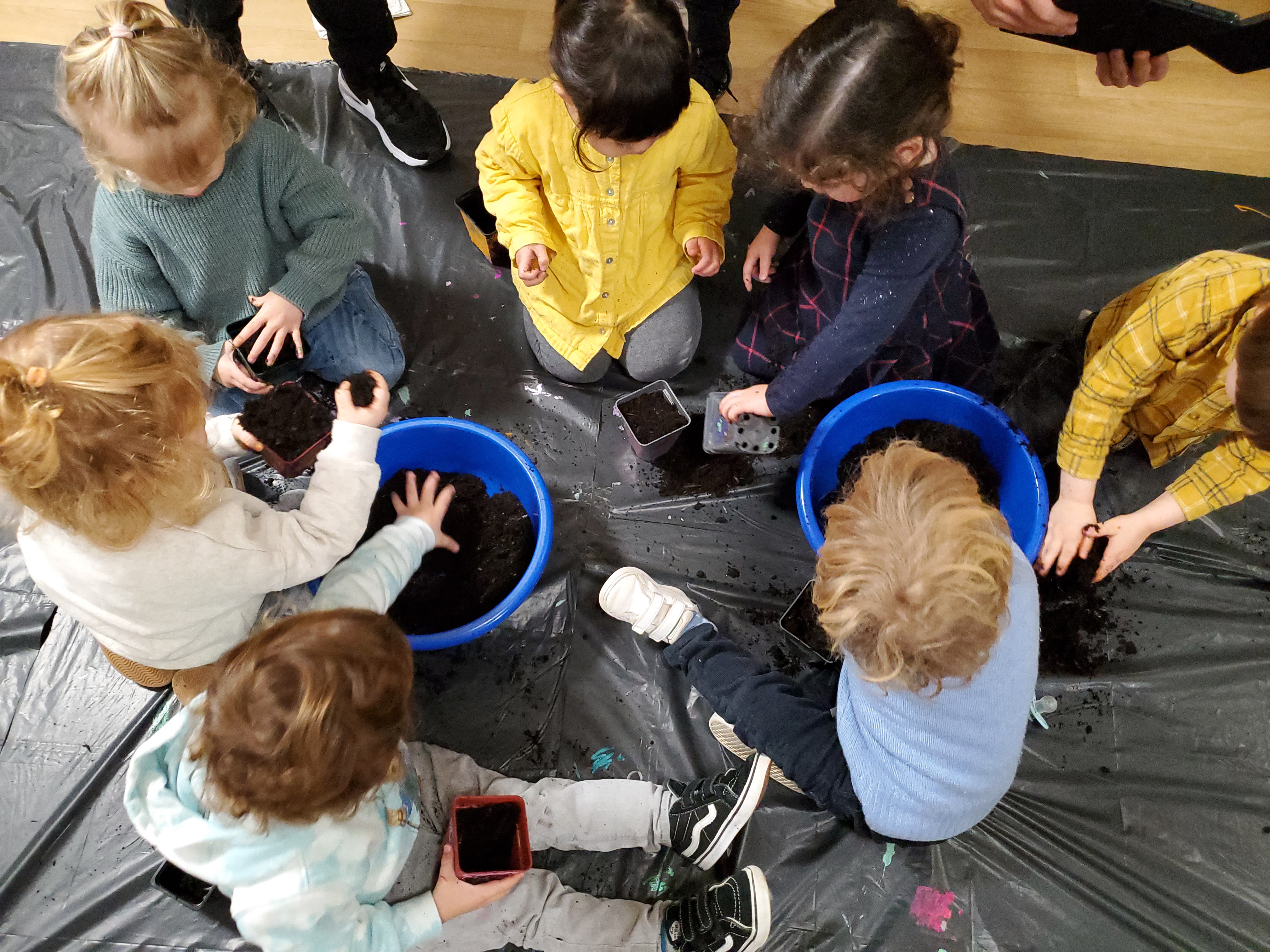 Installés sur une bâche noire autour de deux bassines bleues remplies de terre, un groupe d'enfant remplit des petits pots pour pouvoir planter des graines.