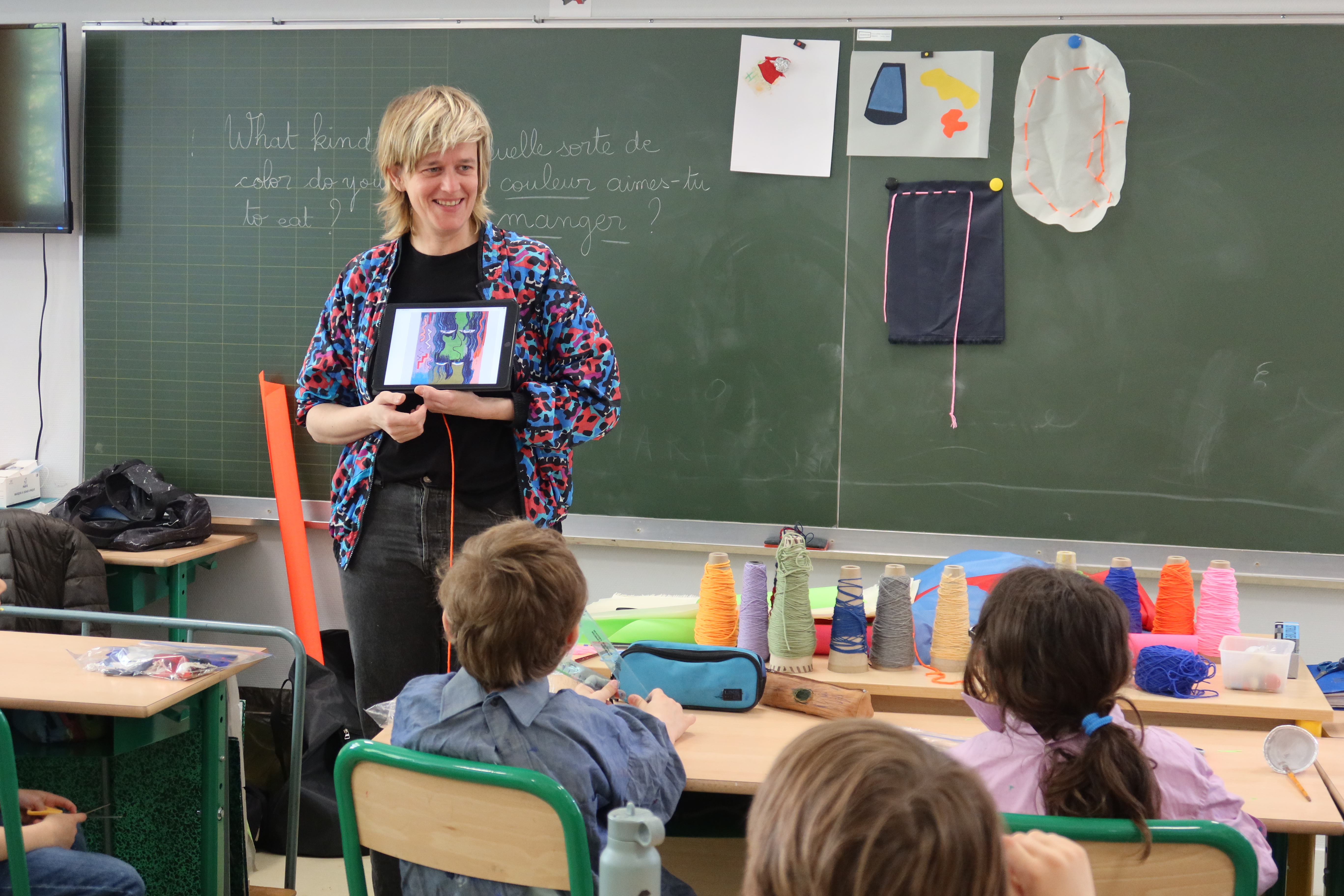 Les élèves sont assis dans leur salle de classe. Face à eux se tient Chloé Dugit-Gros. L'artiste est en train de leur montrer l'écran d'une tablette sur laquelle se trouve des photographies de ses oeuvres. Elle est toute sourire.