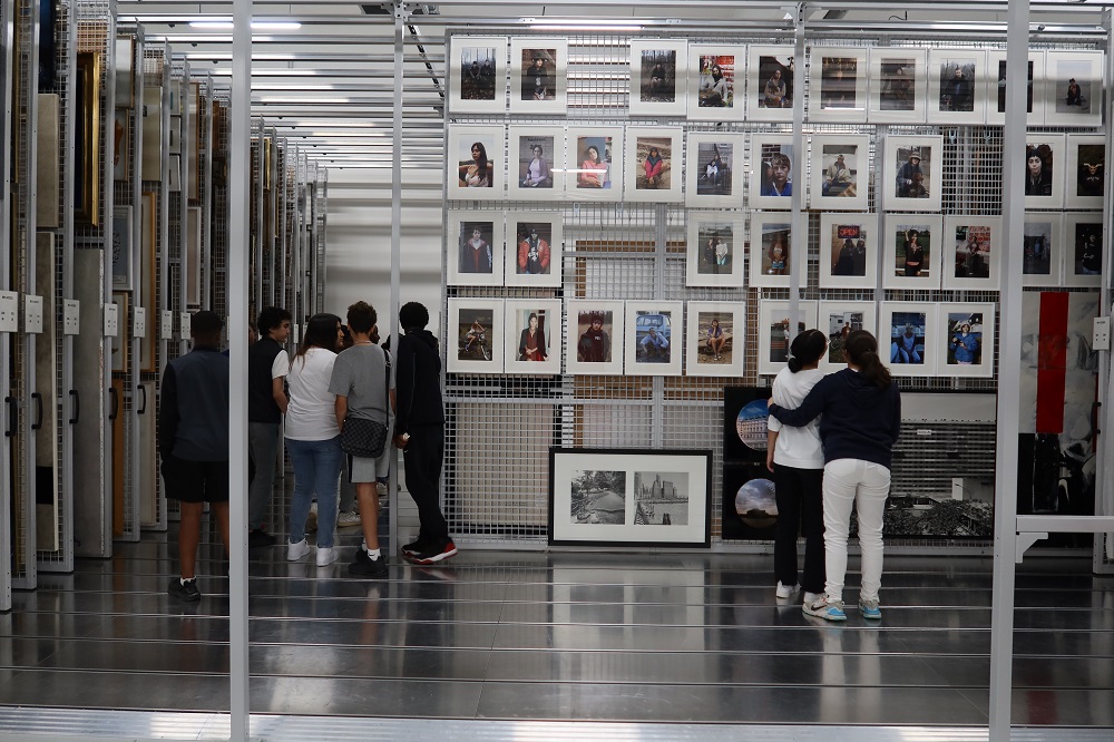 Deux élèves se tiennent devant une grille où sont accrochées les photographies. Un groupe d'élèves est en train de passer derrière la grille sur la gauche.