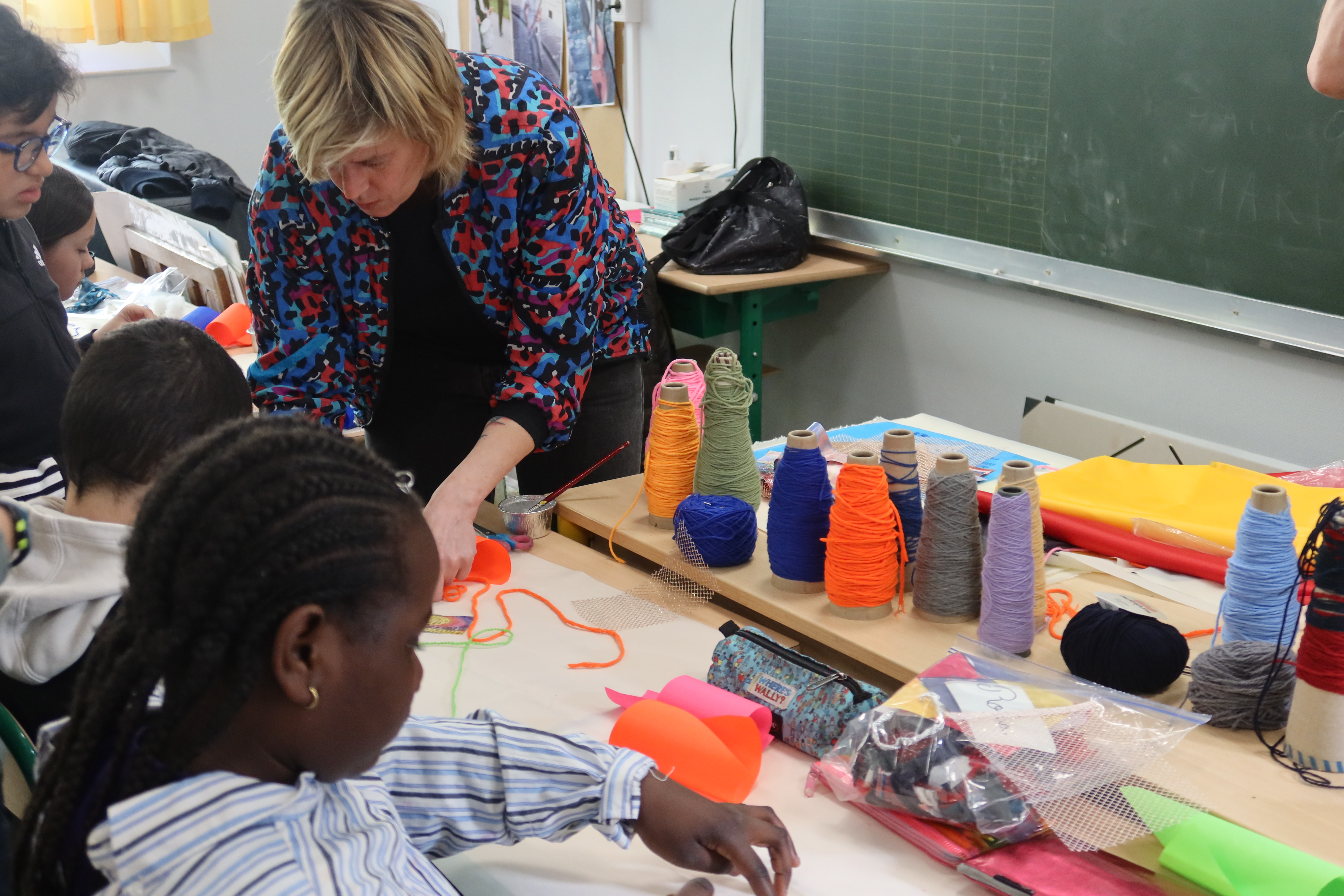 Les élèves sont en train de réaliser un dessin à partir d'éléments textiles disparates pendant que Chloé Dugit-Gros passe les voir pour les aider ou leur expliquer la manière de procéder. L'artiste en penchée sur le pupitre d'un élève