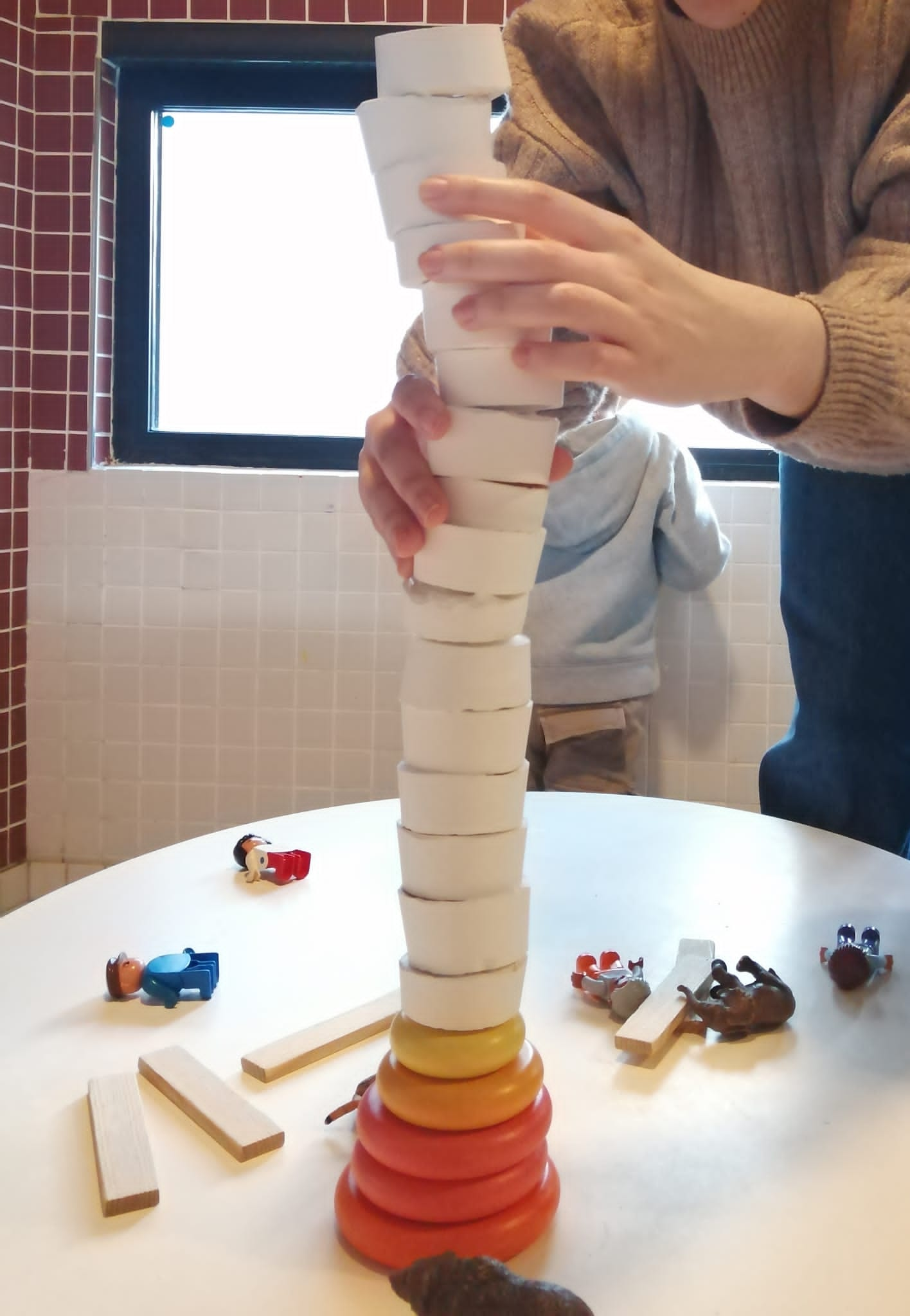 Un adulte aide à faire tenir la plus grande tour de galets de plâtre réalisée par les enfants. La base de la tour est constituée de bloc de construction en forme de galettes : trois oranges de grande taille et de jaune de moyenne taille. Au dessus tiennent en équilibre 16 galets de plâtre.