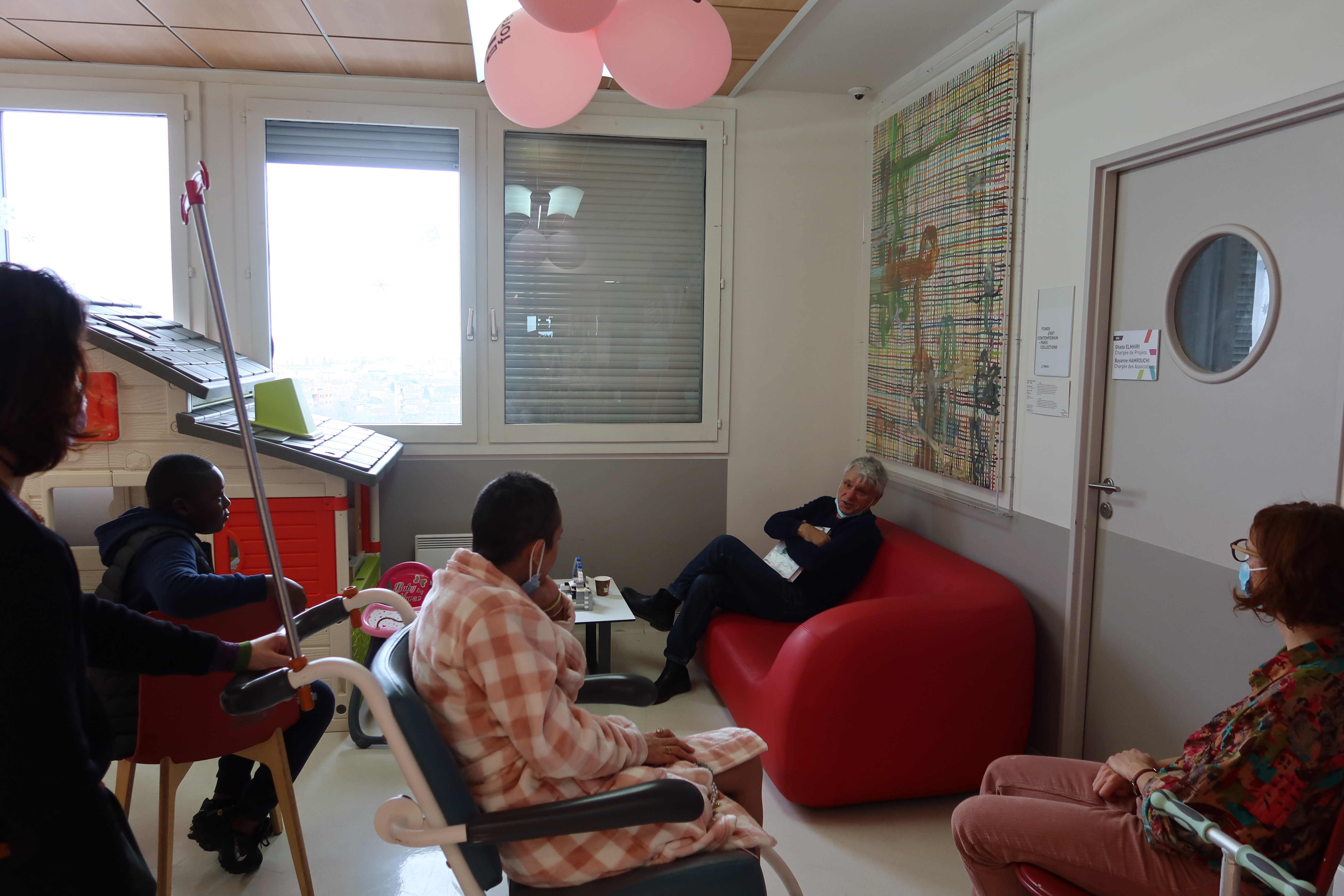 Dans une salle d'attente, l'artiste est assis confortablement dans un canapé rouge. Détendu, il discute avec deux jeunes (un garçon et une femme) qui l'écoute attentivement.