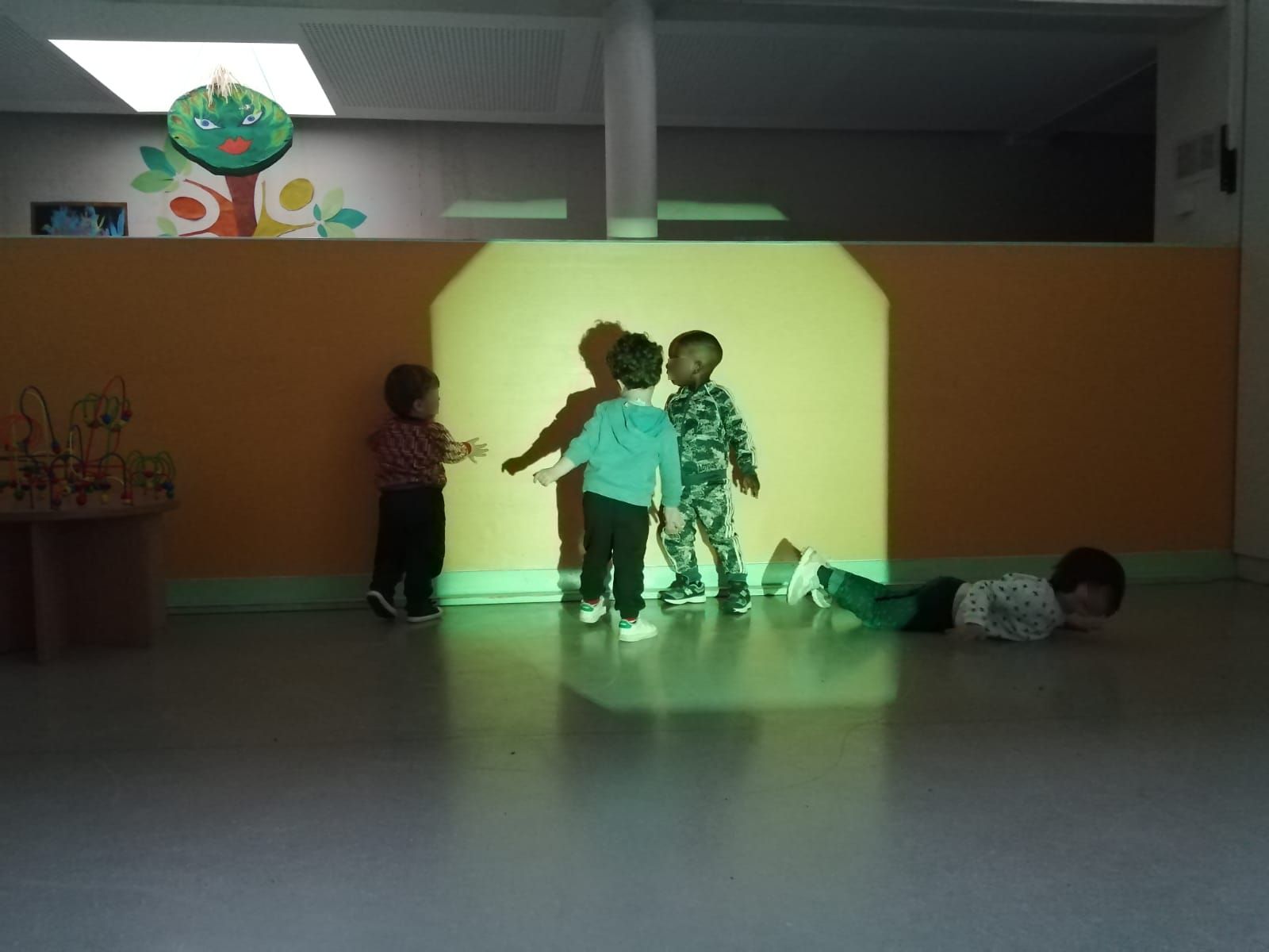 Trois enfants se tiennent face à mur sur lequel de la lumière est projetée. Deux se tiennent dans la lumière et un autre dans l'ombre. Un quatrième enfant est allongé sur le sol, sur le ventre. Il joue lui aussi avec les zones d'ombre et de lumière.