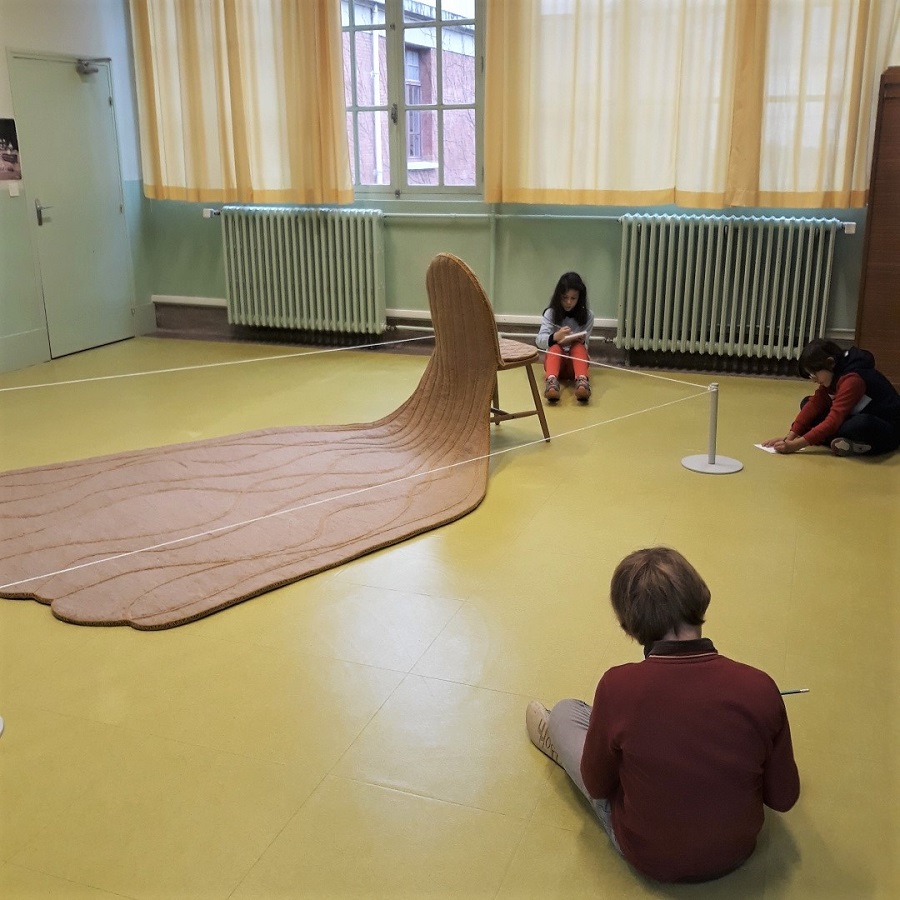 Au sein de leur école, les enfants installés au sol, dessinent l'œuvre, La chaise, de Florence Doléac.