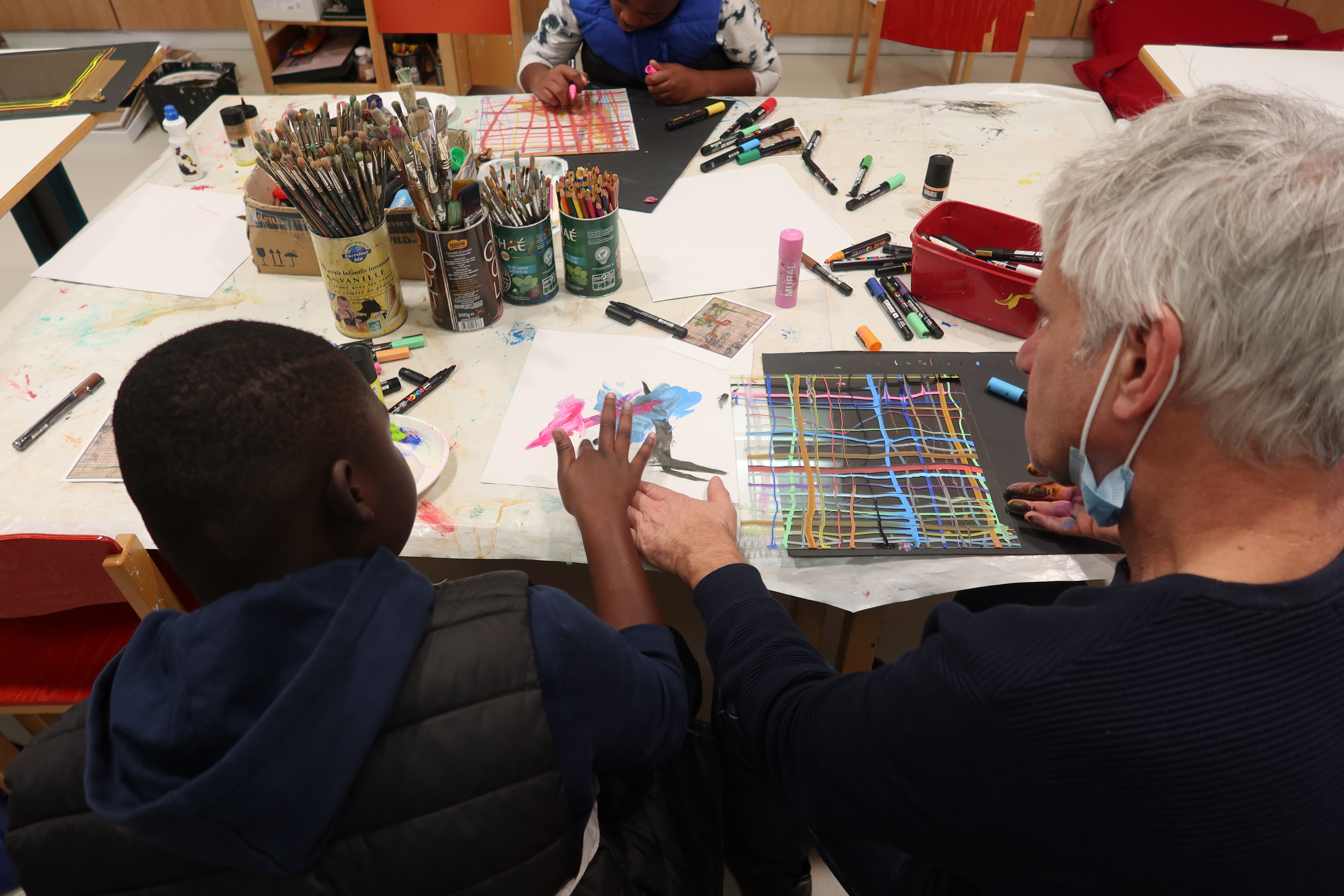 Jérôme Boutterin et un garçon du centre hospitalier sont en train d'étaler la peinture à la main afin de créer des tâches de peinture abstraite. L'artiste et l'enfant travaillent ensemble. Il lui montre sa manière de faire et lui explique comment procédé.